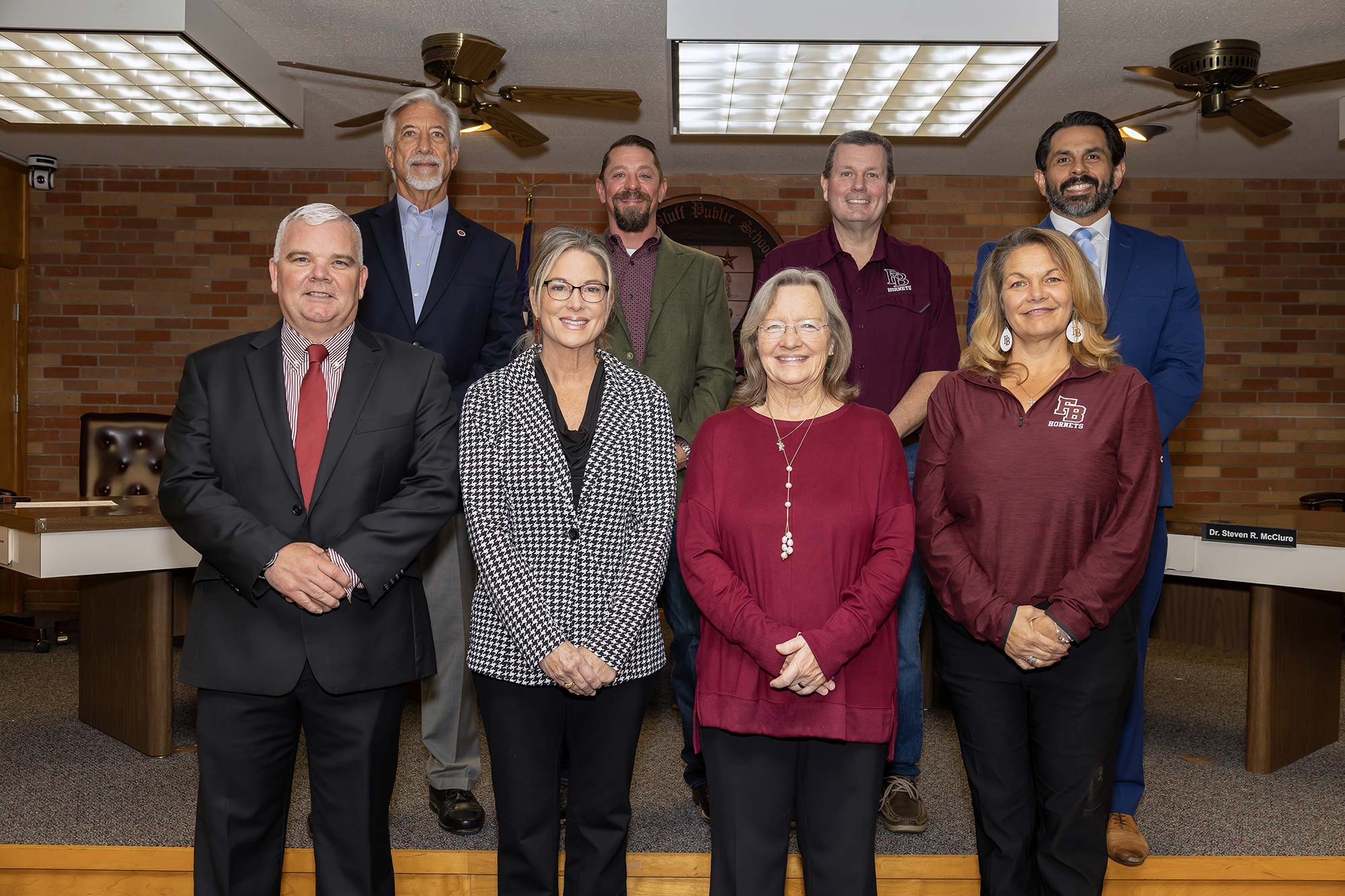 Eight adults pose and smile for a group photo in a meeting room. Four people stand in the front row and four in the back. They wear business or business-casual attire, with desks and brick walls visible in the background.