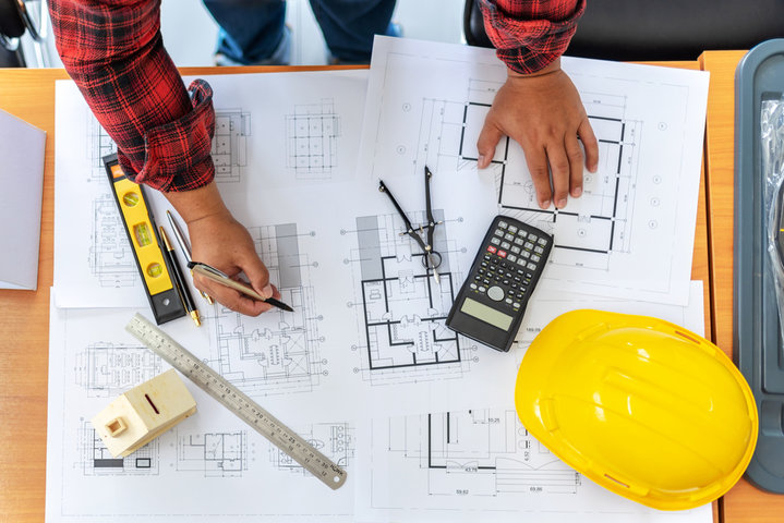 A person in a red plaid shirt reviews blueprints on a desk, holding a pen. Nearby are a calculator, compass, level, ruler, measuring tape, and a yellow construction hard hat.