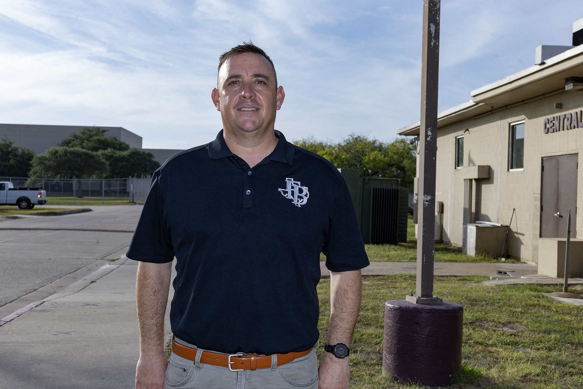 A man wearing a navy polo shirt and khaki pants stands outdoors on a sidewalk near a beige building with CENTRAL on the door, under a partly cloudy sky.