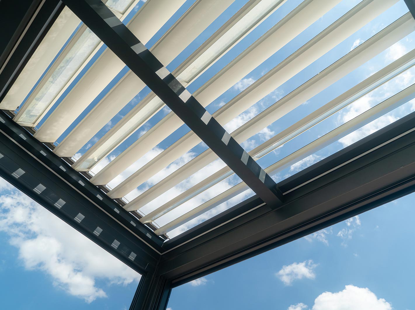 View looking up through a roof with adjustable slats, partially open to reveal a bright blue sky with scattered white clouds. The slats create geometric shadows and lines.