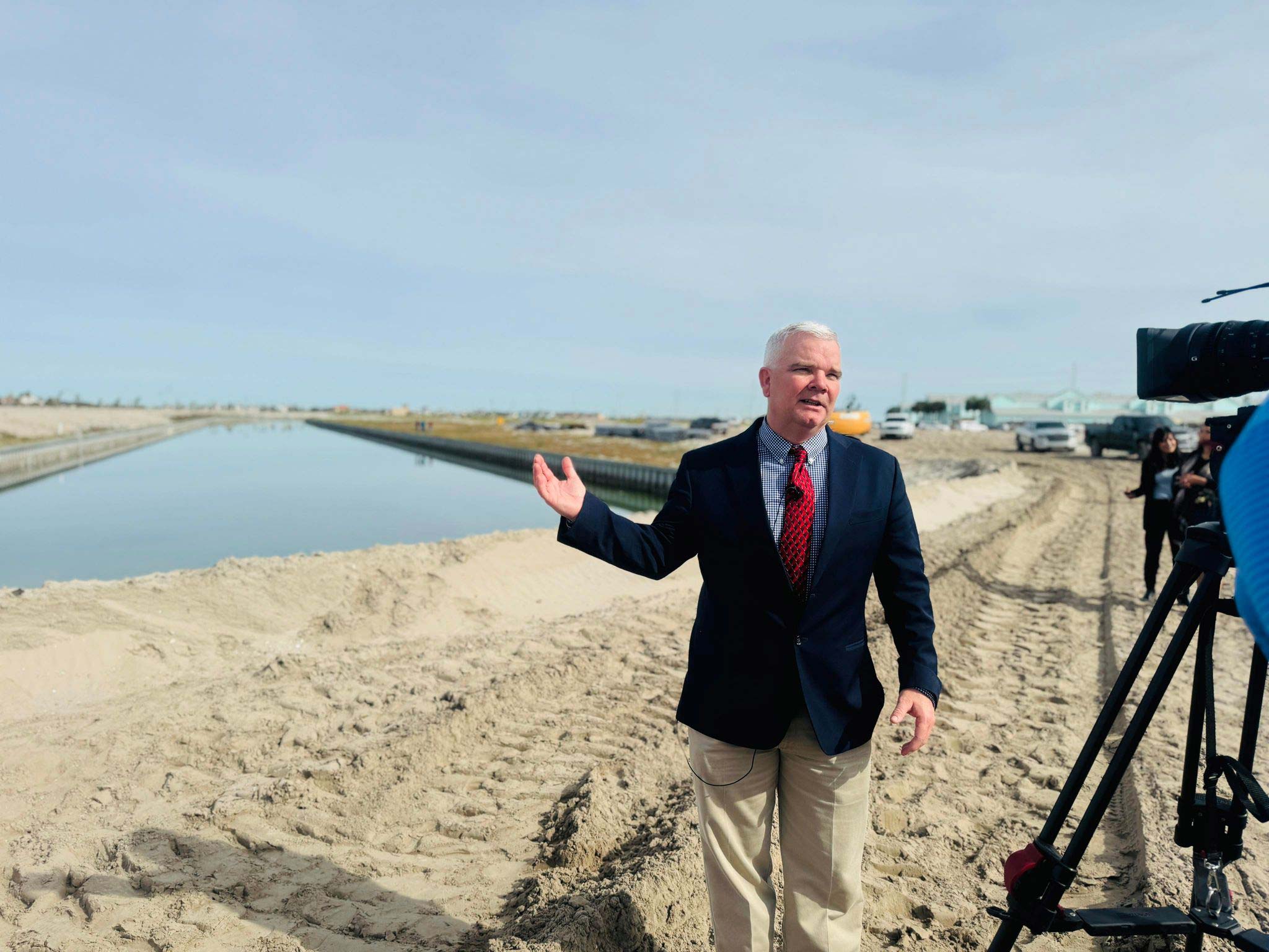 A man in a suit gestures while standing on a sandy construction site next to a canal, with a camera filming him and vehicles and structures in the background under a partly cloudy sky.
