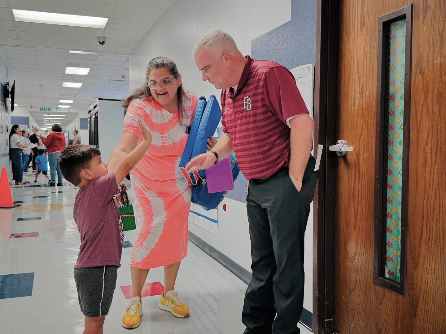 A young boy gives a fist bump to a smiling man in a maroon shirt as a woman in a pink dress stands beside him in a school hallway. Other people are visible in the background.