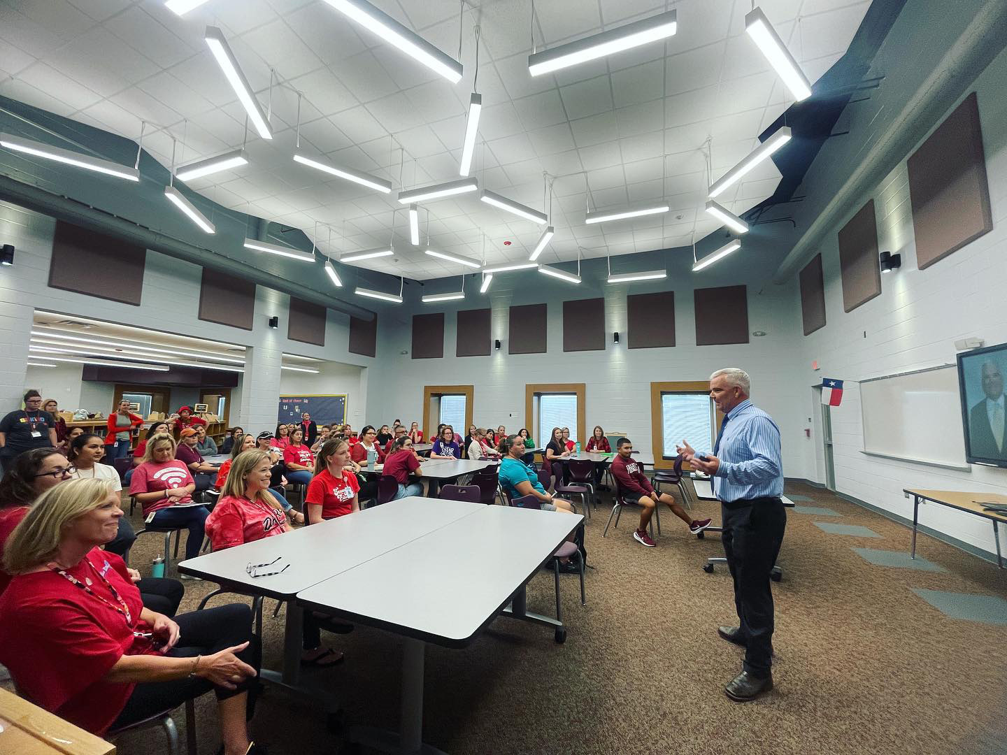 A man in a blue shirt speaks to a group of people seated at tables in a bright, modern classroom with geometric ceiling lights. Most attendees wear red shirts and listen attentively. A screen displays another man remotely.