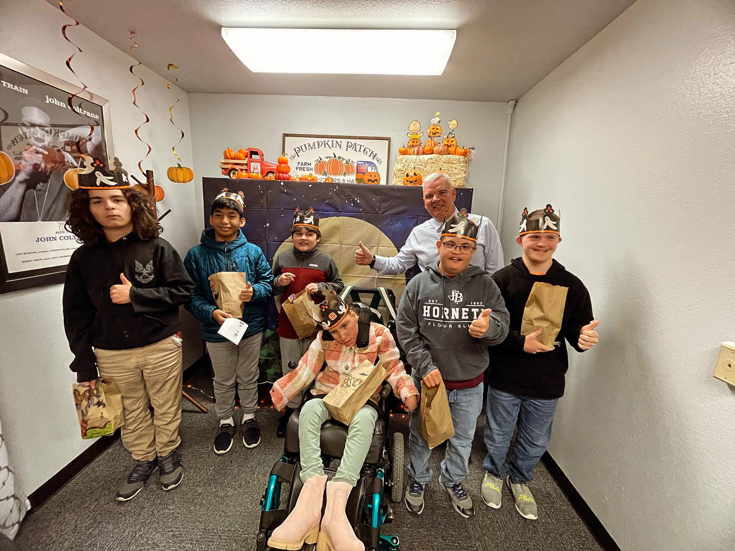 A group of six smiling kids, one in a wheelchair, and an adult stand in a decorated room with pumpkin-themed decor. All wear Burger King crowns and hold brown bags, giving thumbs up to the camera.