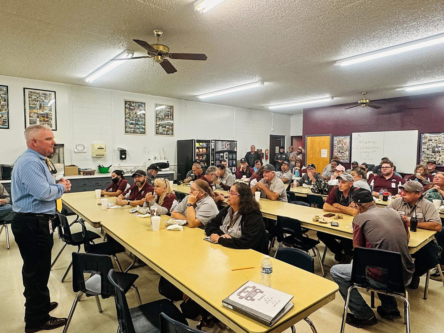 A man stands at the front of a crowded meeting room, speaking to a group of seated people at long yellow tables. Attendees listen attentively, some eating, with papers and drinks on the tables.