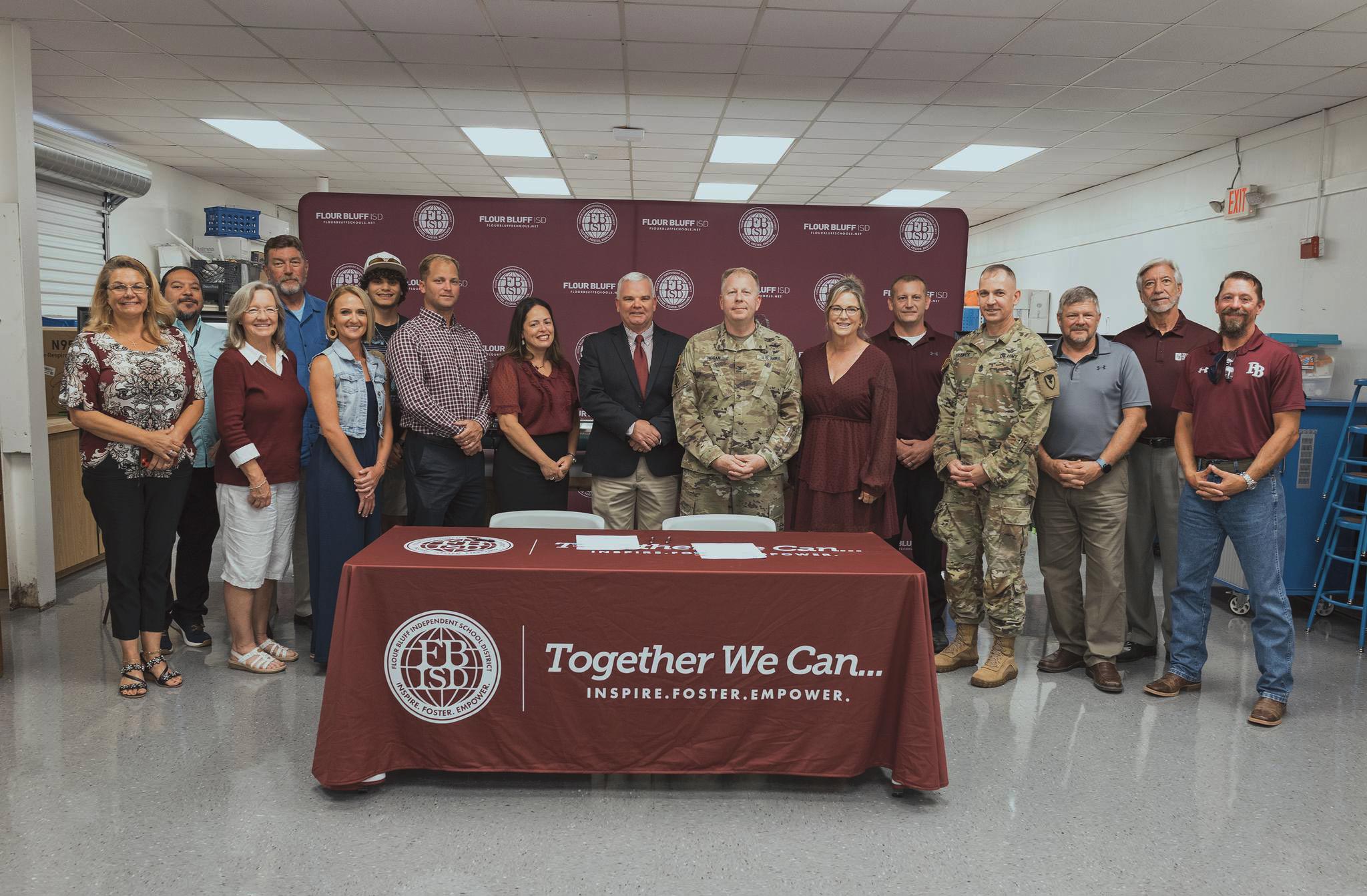 A group of people, including civilians and military personnel, stand behind a table with a maroon cloth displaying the slogan Together We Can... Inspire. Foster. Empower. and the FBISD logo, in a well-lit room.