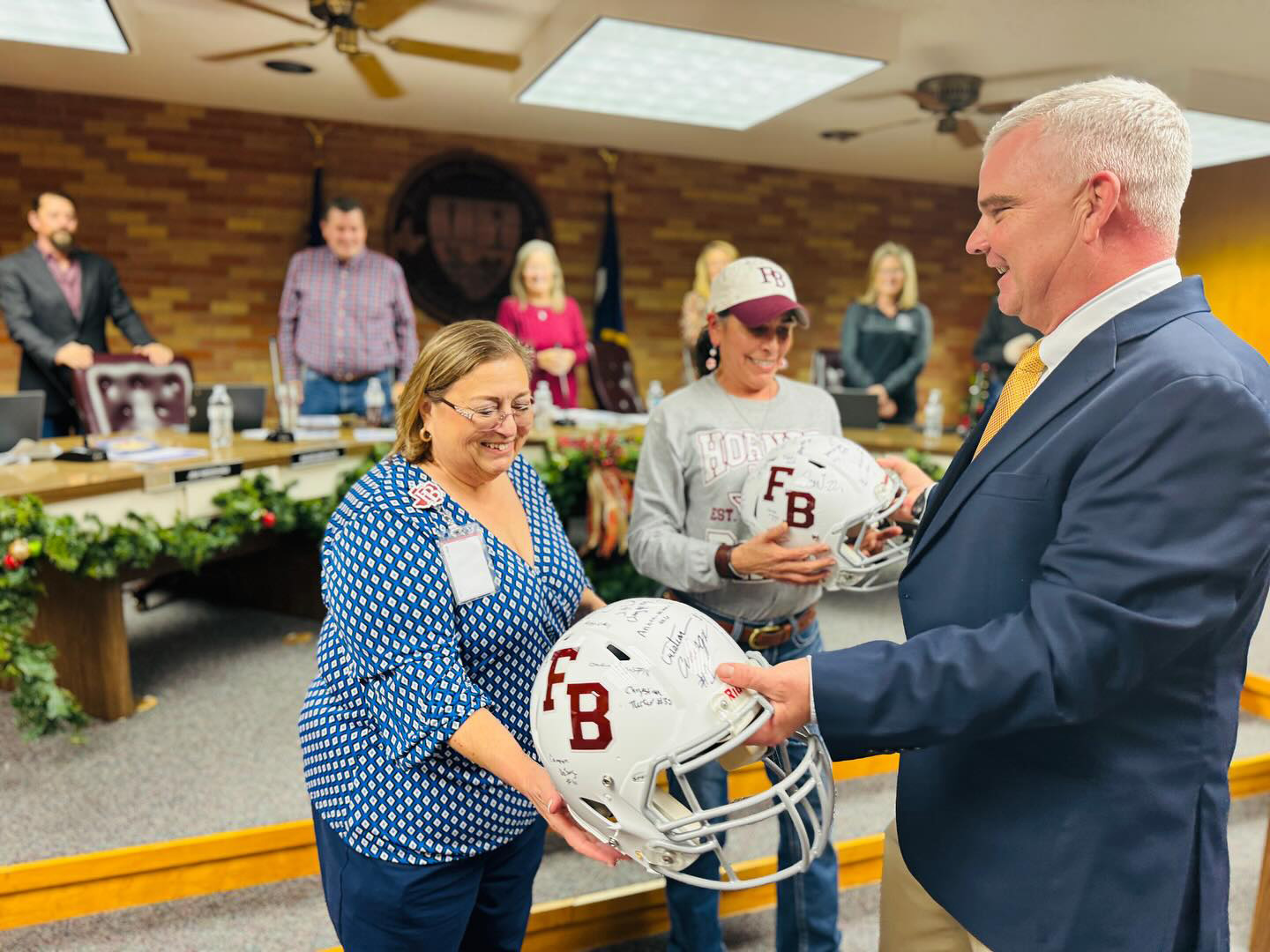 A man in a suit hands a signed football helmet to a smiling woman while others watch in a decorated room with a brick wall and holiday greenery. Two women hold FB helmets and appear happy during the presentation.