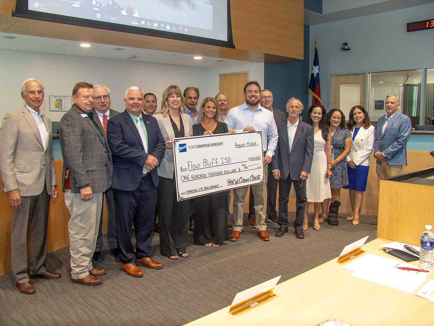 A group of people stands together indoors, smiling at the camera and holding an oversized check for $100,000 made out to Flour Bluff ISD. A Texas flag is visible in the background.