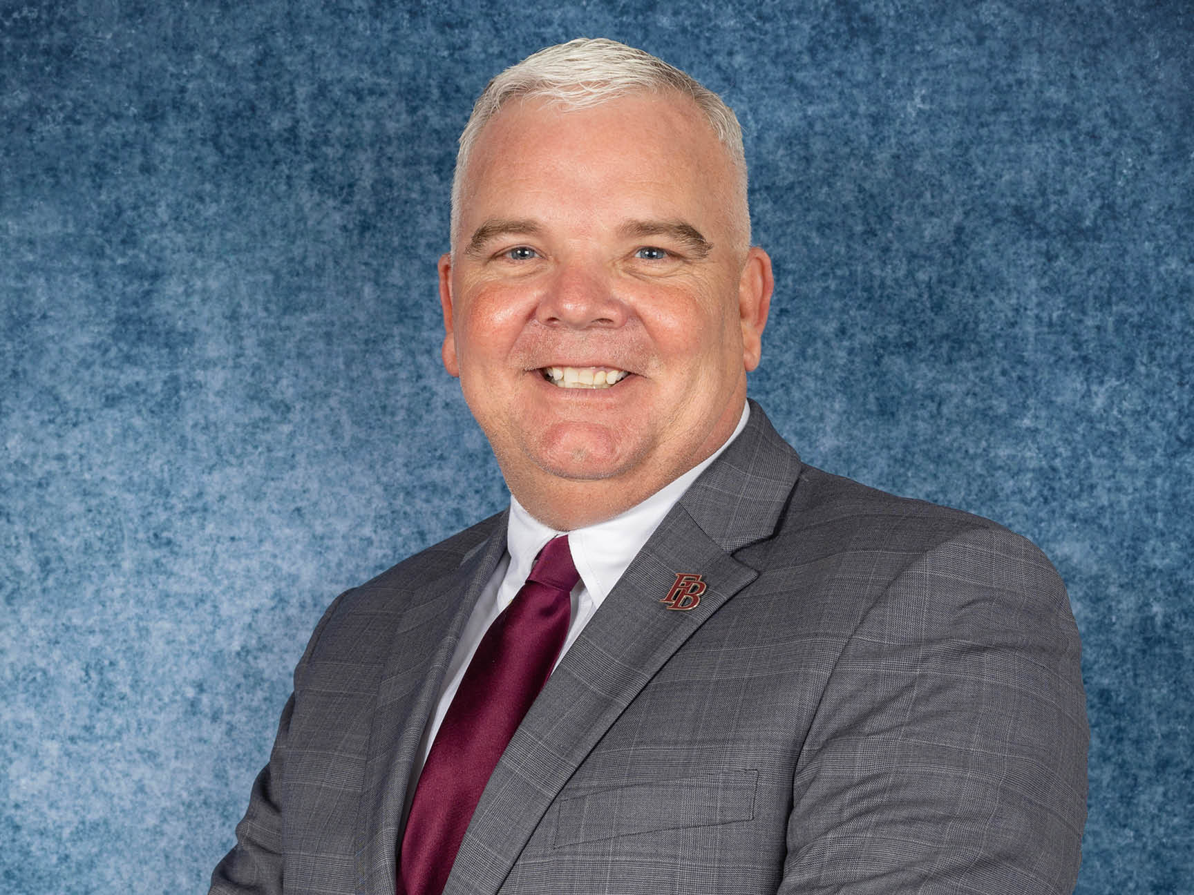 A smiling man with short white hair wearing a gray checked suit, maroon tie, and a pin on his lapel, posed in front of a blue textured background.