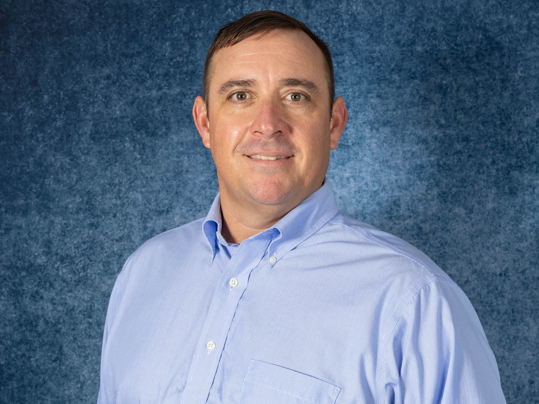 A man with short brown hair wearing a light blue button-up shirt smiles at the camera in front of a textured blue background.