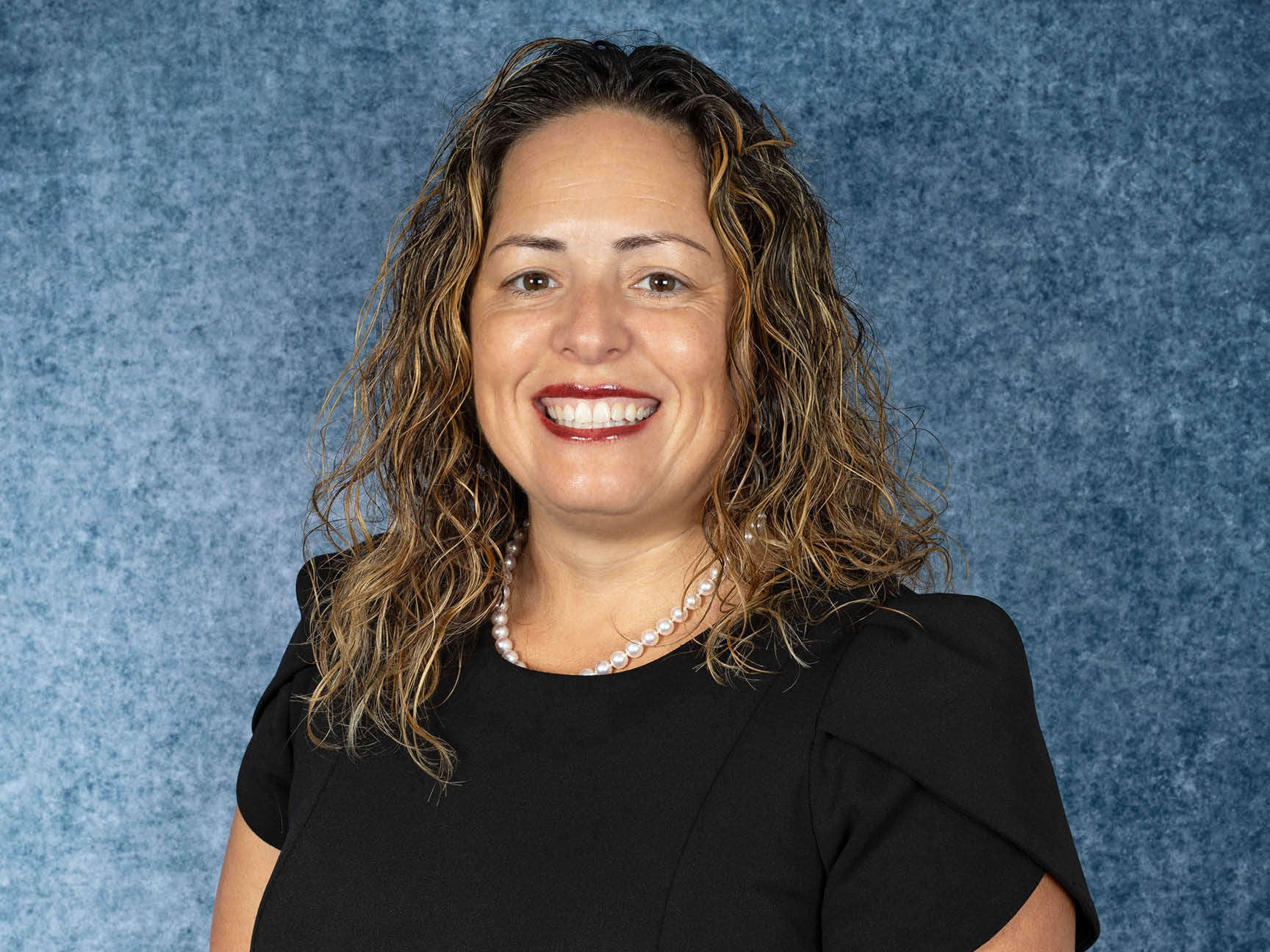 A woman with curly brown hair, wearing a black dress and a pearl necklace, smiles at the camera against a mottled blue background.