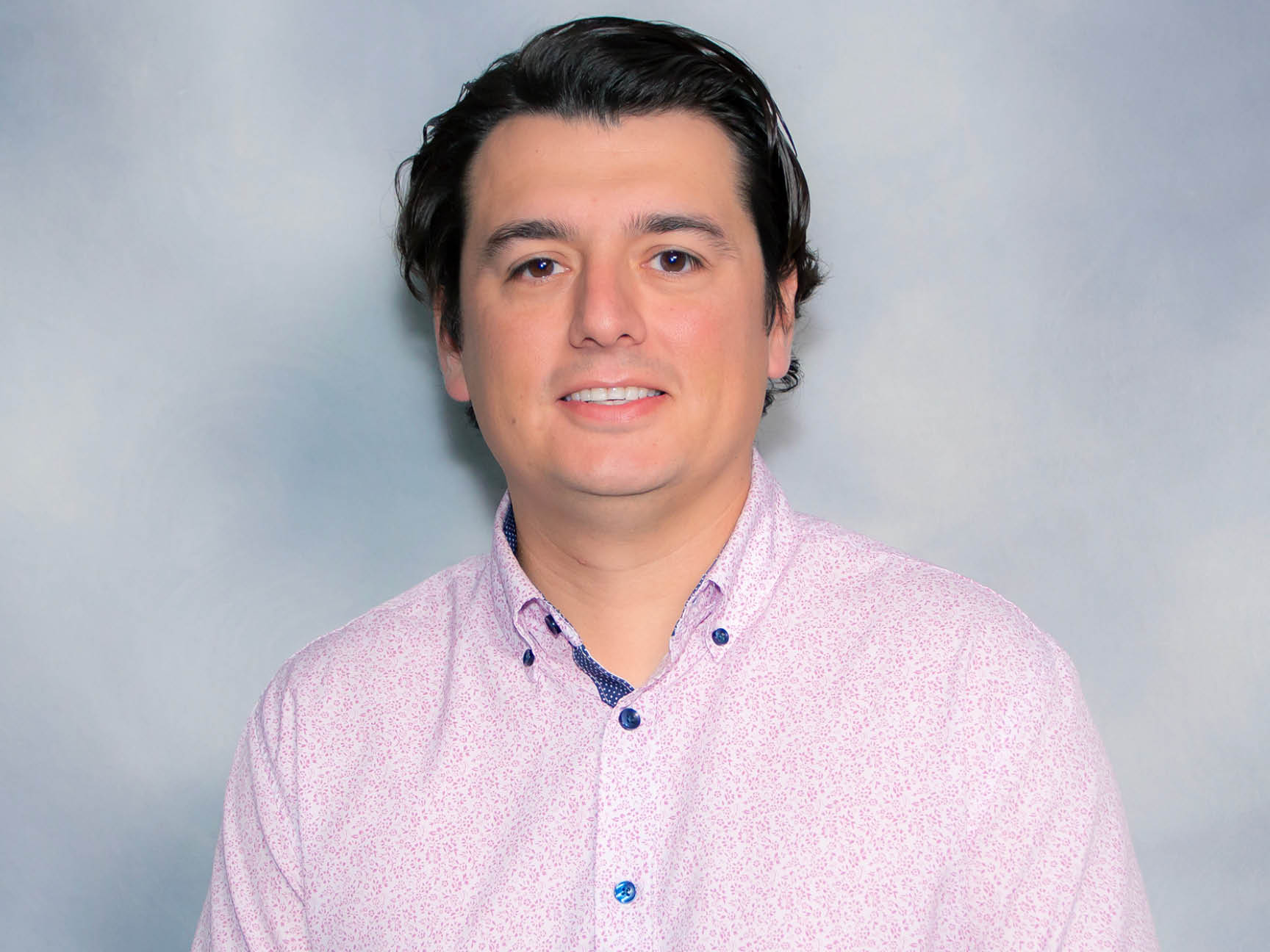 A man with short dark hair, wearing a light pink patterned button-up shirt, stands in front of a soft gray and blue gradient background, smiling slightly at the camera.