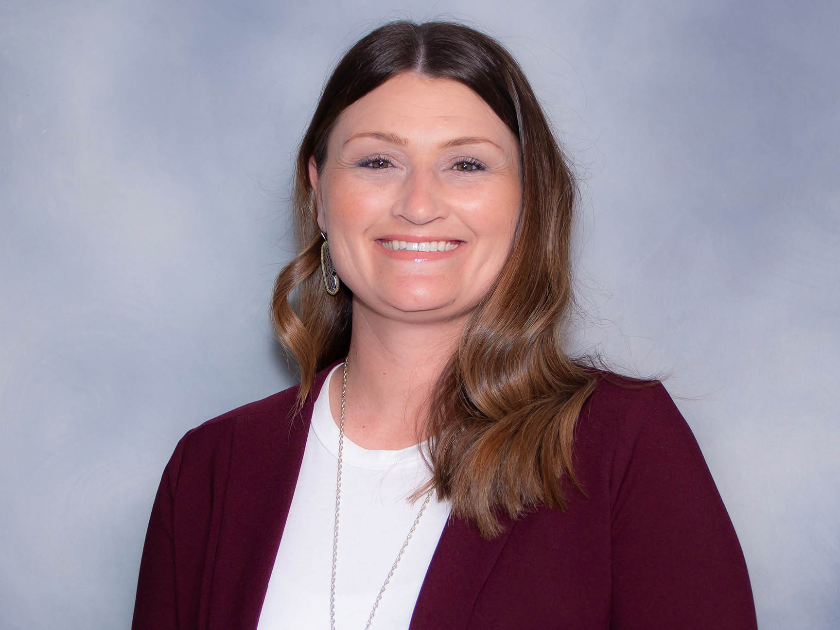 A woman with long brown hair, wearing a maroon blazer over a white top and a necklace, smiles in front of a light gray background.