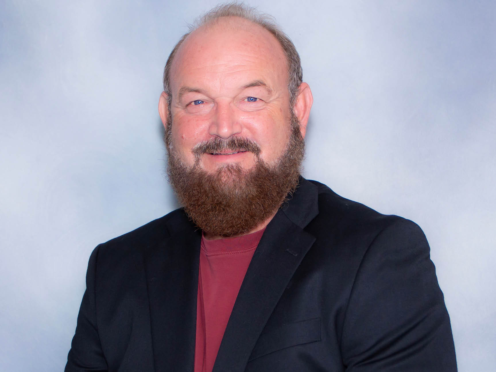 A middle-aged man with a bald head and full brown beard, wearing a black blazer over a maroon shirt, smiles in front of a light gray background.