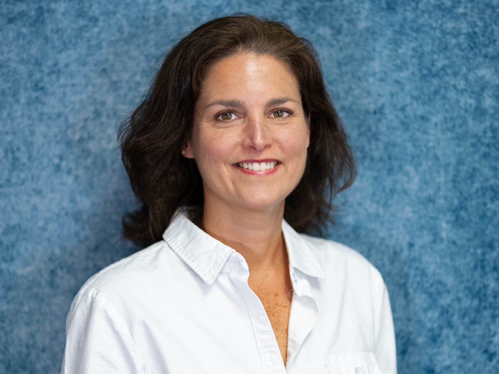 A woman with shoulder-length brown hair and a white button-up shirt smiles in front of a textured blue background.
