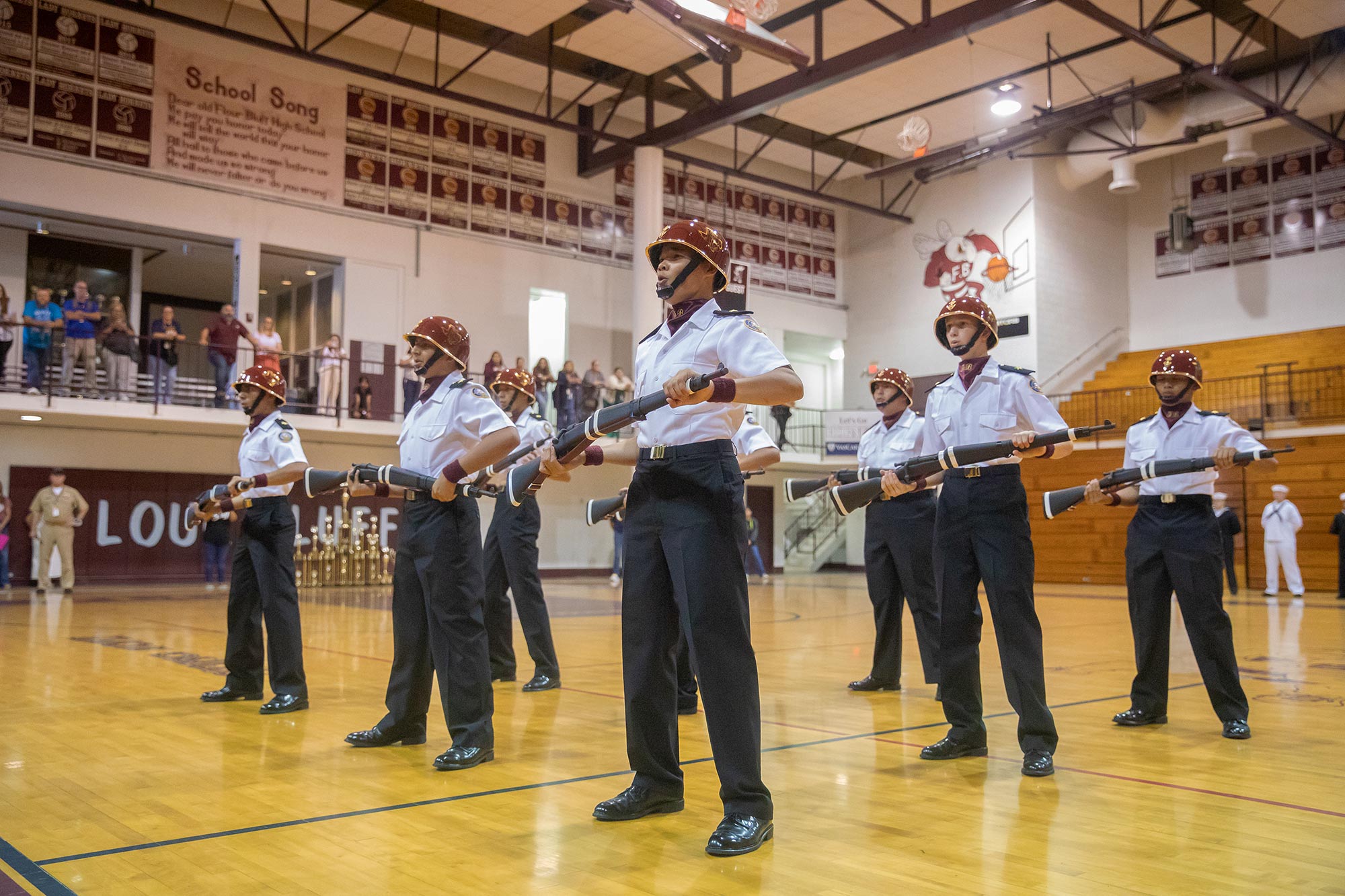 A group of uniformed cadets in helmets perform a synchronized rifle drill on a gymnasium floor, watched by spectators from bleachers and a balcony. Banners and school mascot art are visible on the walls.