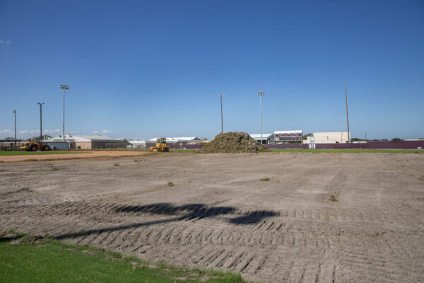 A baseball field under renovation with exposed dirt, construction vehicles, and a large pile of soil in the center; buildings and a scoreboard are visible in the background under a clear blue sky.