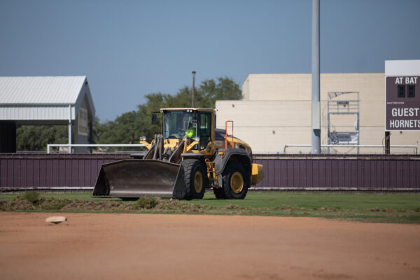 A person operating a yellow front-end loader is clearing dirt on a baseball field. Buildings, a scoreboard, and a fence are visible in the background under a clear blue sky.