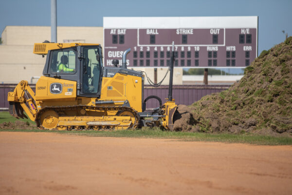A yellow bulldozer moves dirt on a baseball field, with a large scoreboard and a pile of soil in the background.