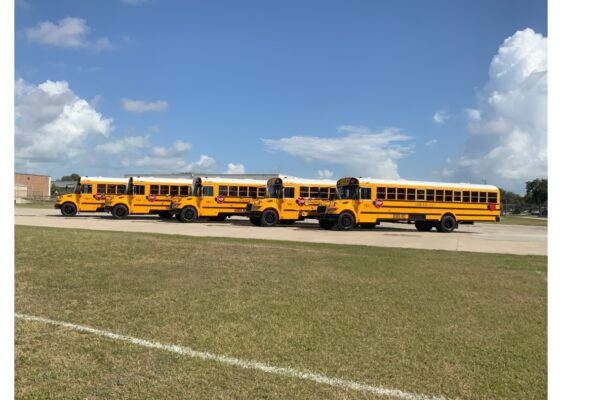 Four yellow school buses are parked in a row on a paved lot next to a grassy field, under a partly cloudy blue sky. A brick building can be seen in the background on the left.