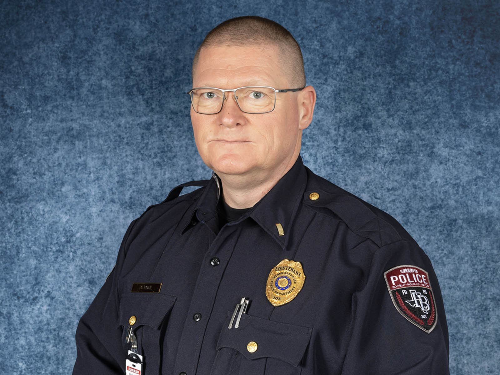 A police officer wearing glasses and a dark uniform with badges, pins, and an ID card poses in front of a mottled blue backdrop.