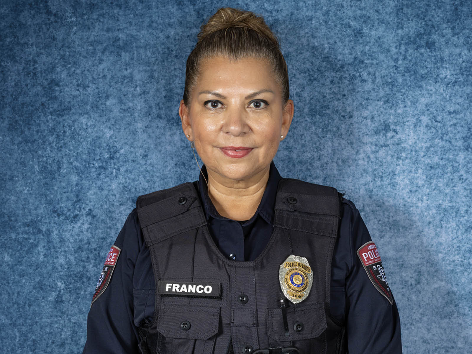A police officer with light brown hair in a bun, wearing a dark uniform and bulletproof vest labeled Franco, stands in front of a blue textured background, facing the camera and smiling slightly.