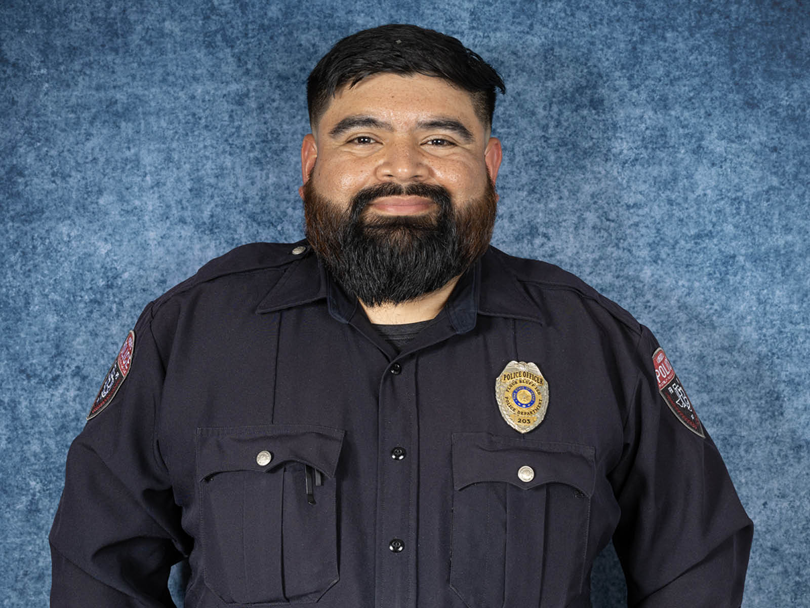 A man with a beard wearing a dark police uniform and badge stands smiling in front of a mottled blue backdrop.