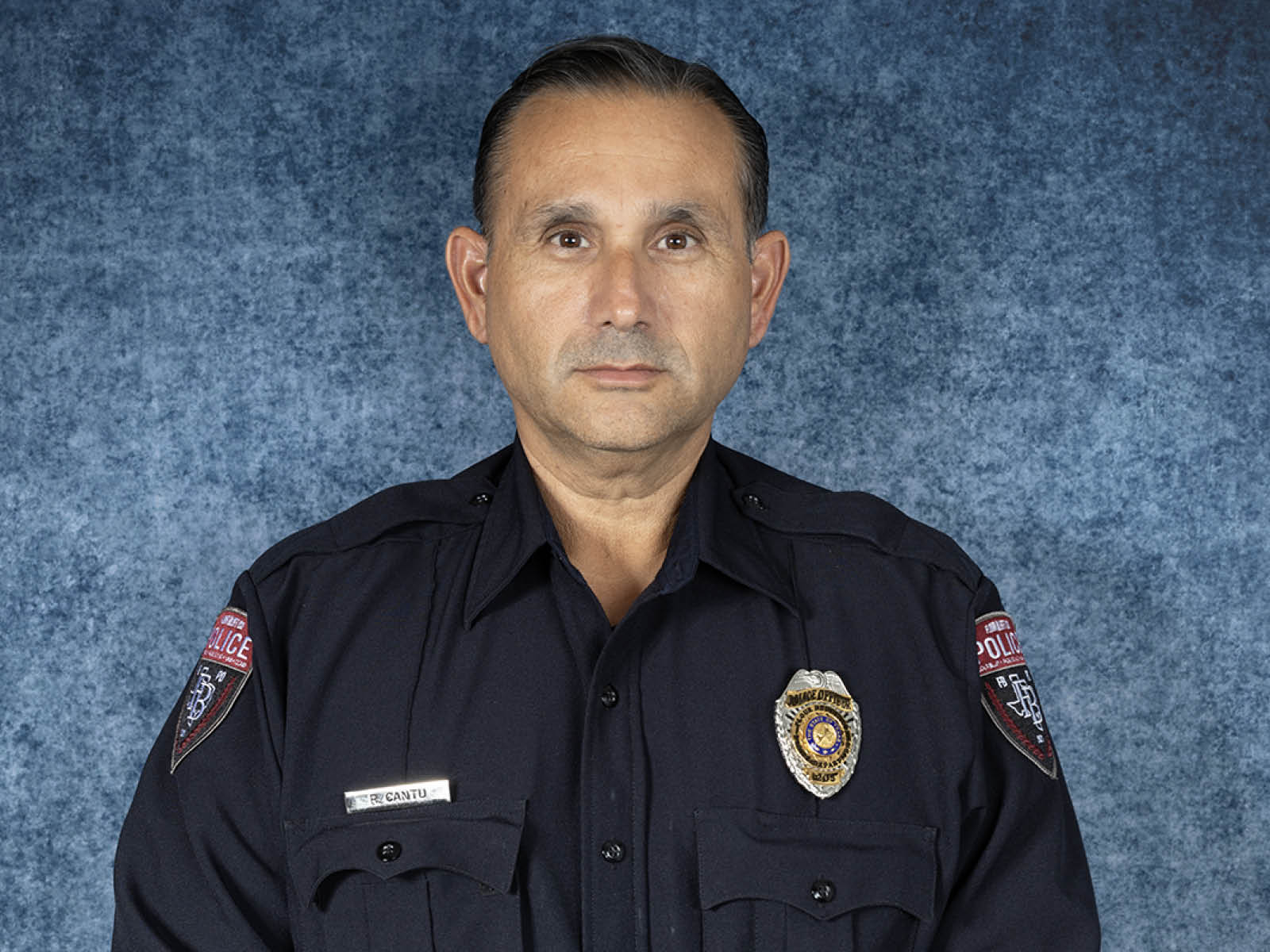 A man in a dark police uniform with badges and name tag poses against a mottled blue background for a formal portrait.