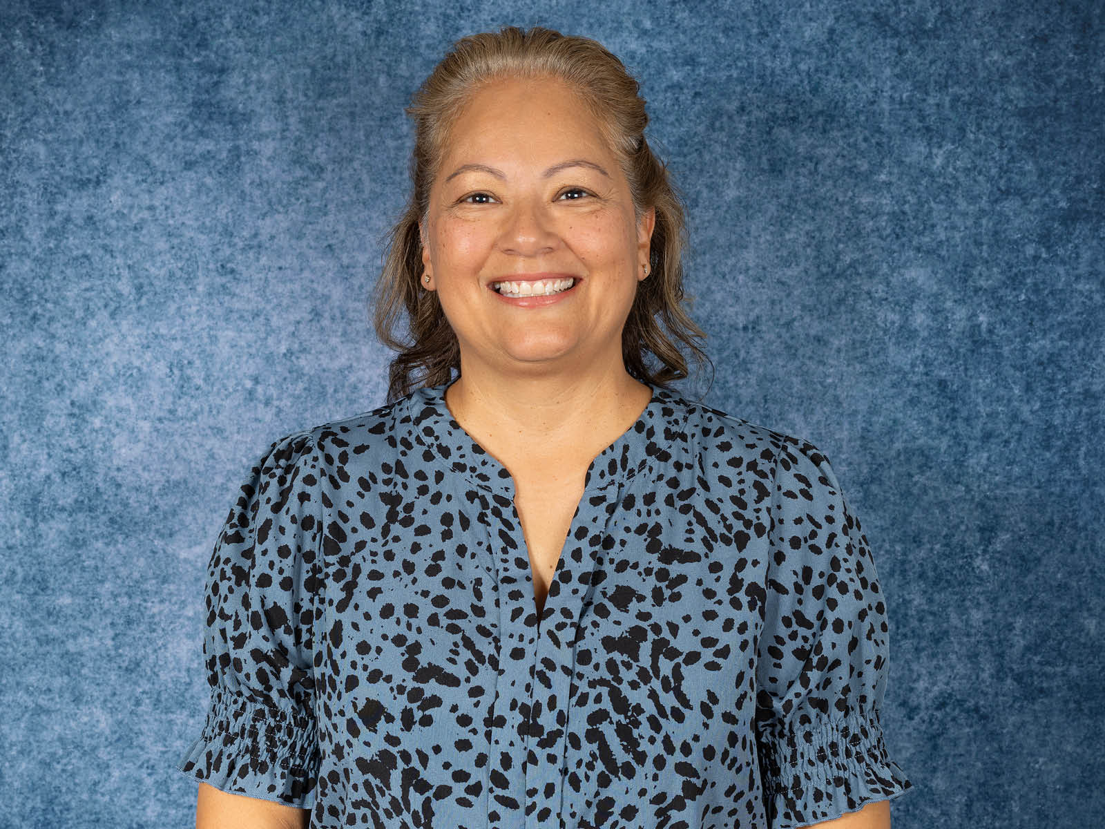 A smiling woman with light brown hair wearing a blue blouse with a black spotted pattern stands in front of a textured blue background.