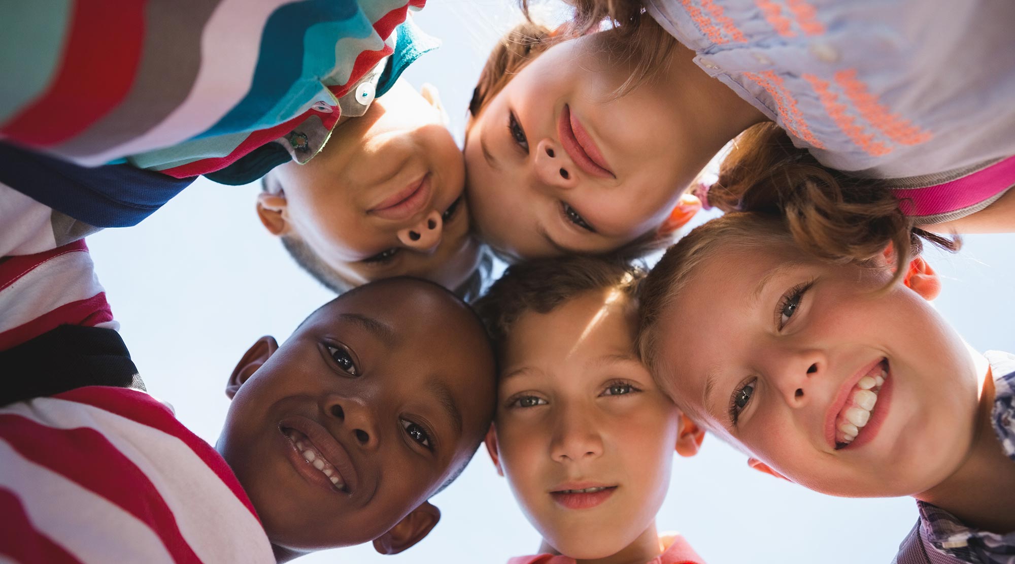 A group of five smiling children stand in a circle, facing inward and looking down at the camera, with a clear blue sky in the background.