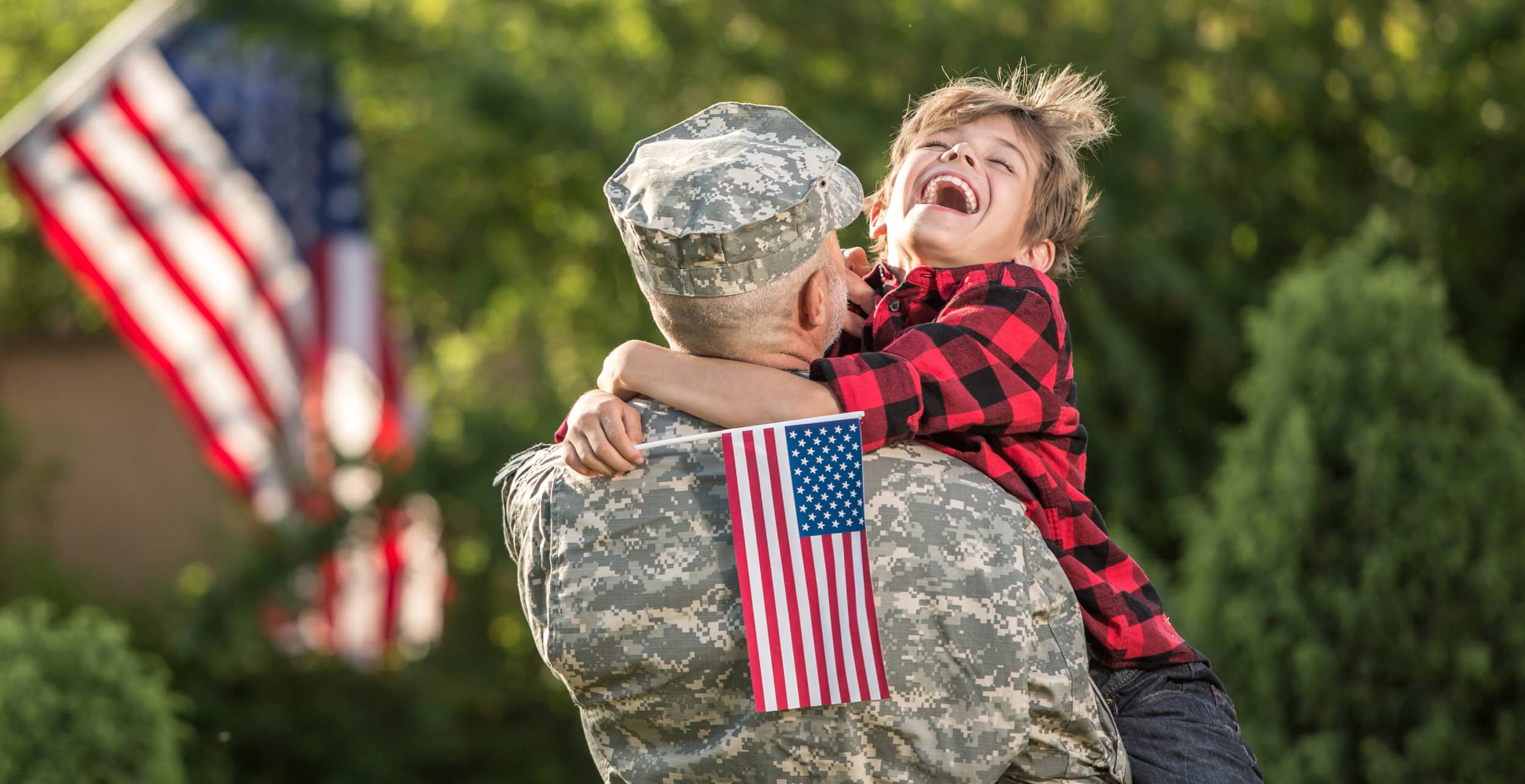 A smiling child in a red plaid shirt joyfully hugs a person in military uniform holding a small American flag outdoors, with a larger flag blurred in the background.
