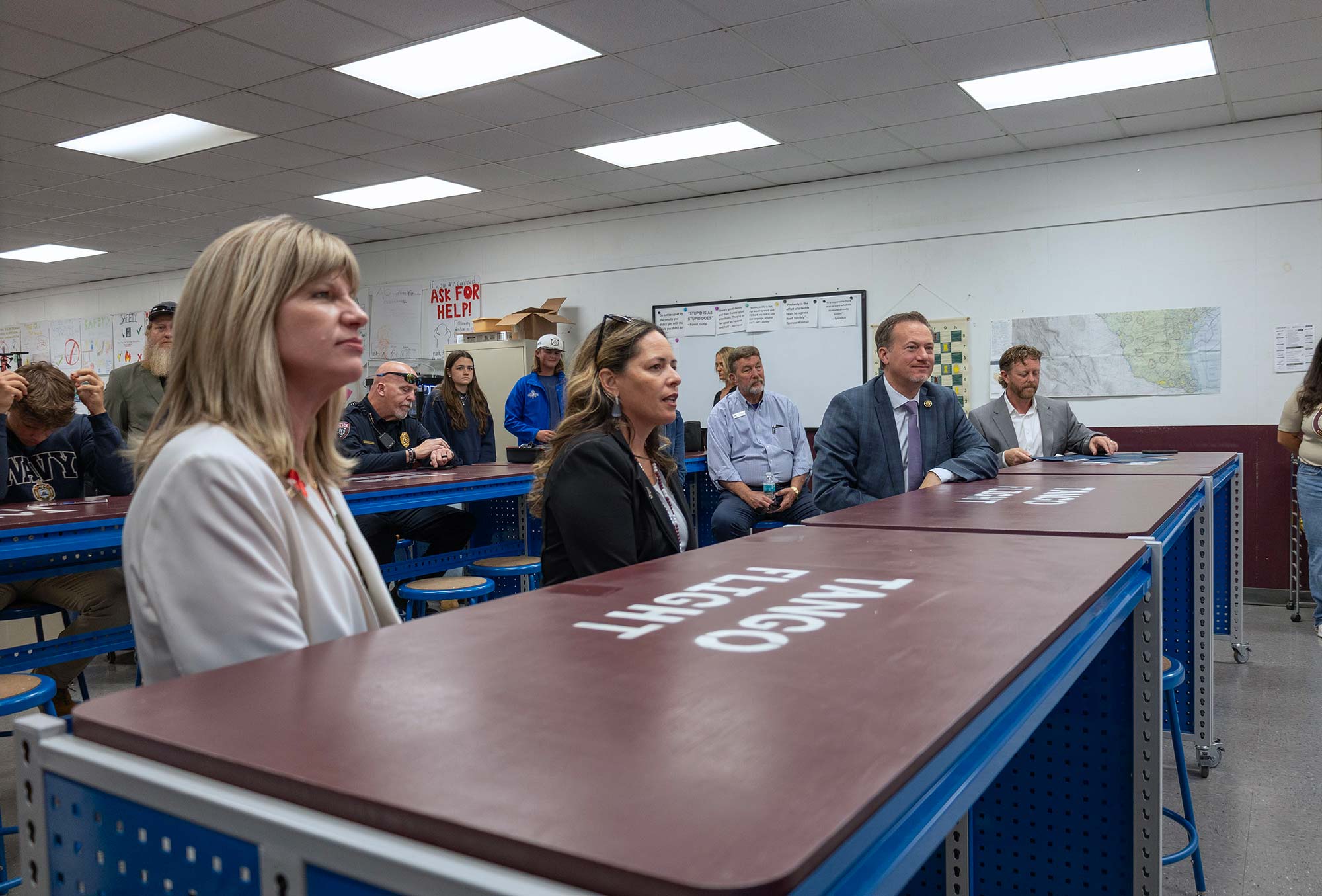 A group of adults sit at blue tables in a classroom, facing forward. Three people sit at the front with “Tango” and “Flight” written on the tables, while others sit and watch in the background. Maps and posters hang on the walls.