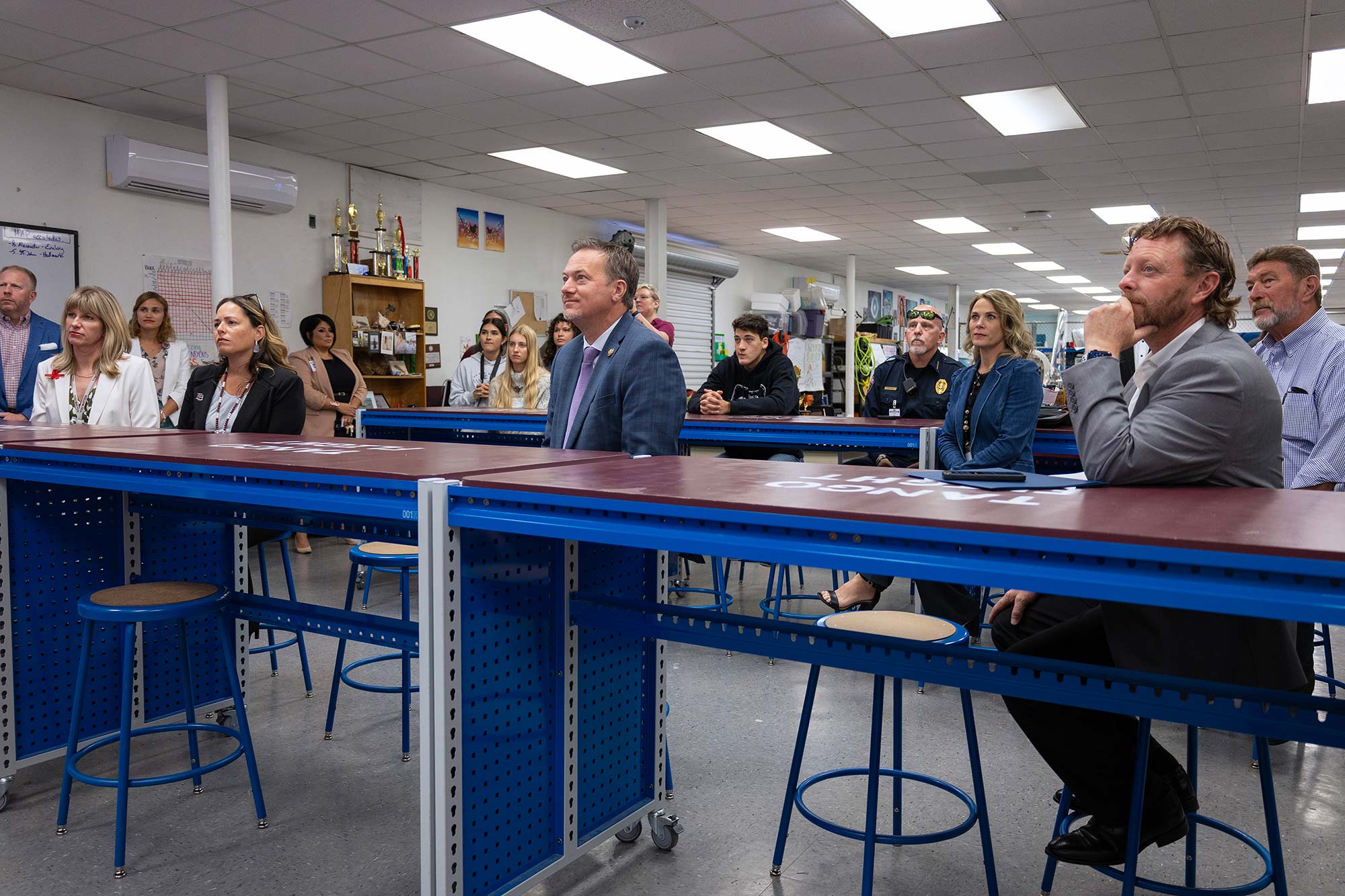 A group of people, both men and women, sit attentively at blue tables in a brightly lit classroom or lab, listening to someone off-camera. Shelves and trophies are visible in the background.