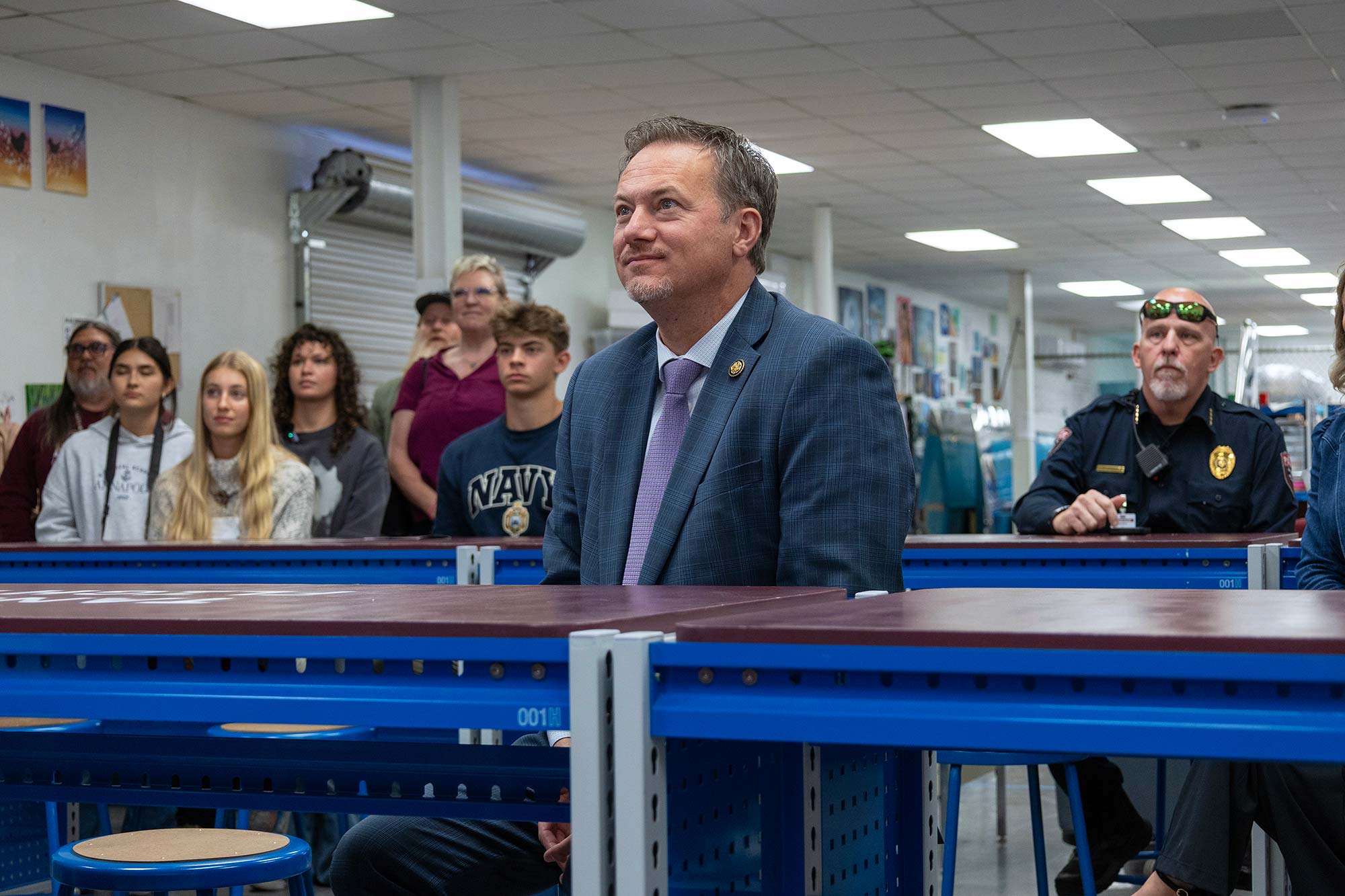 A man in a blue suit and tie sits in a classroom with blue tables, facing forward. Several people, including students and a police officer, are seated and standing in the background. The setting appears educational.