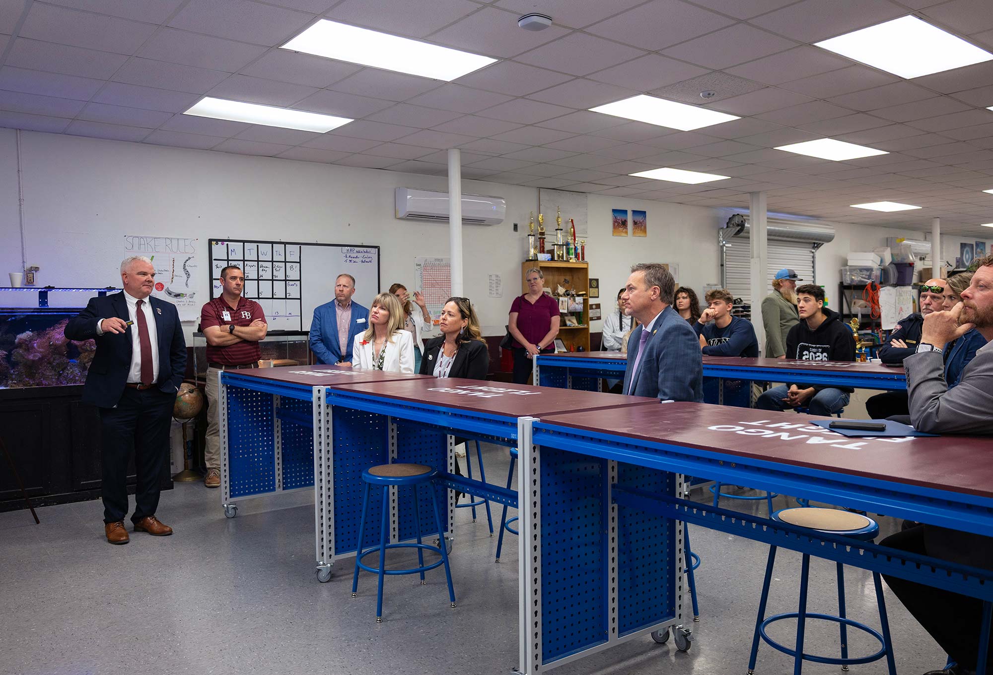 A man in a suit speaks to a group of people seated and standing around blue workbenches in a classroom or lab setting. Aquariums, supplies, and a whiteboard are visible in the background.