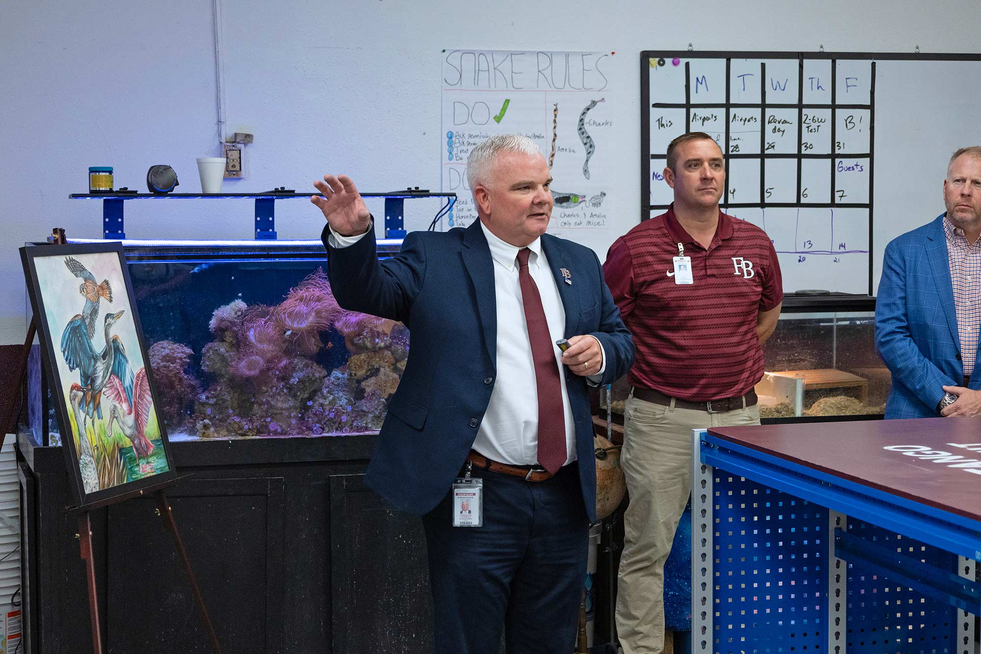 A man in a suit speaks and gestures in a classroom or lab, standing near a fish tank and a colorful painting, with two other men beside him. Behind them are a whiteboard and a classroom rules poster.