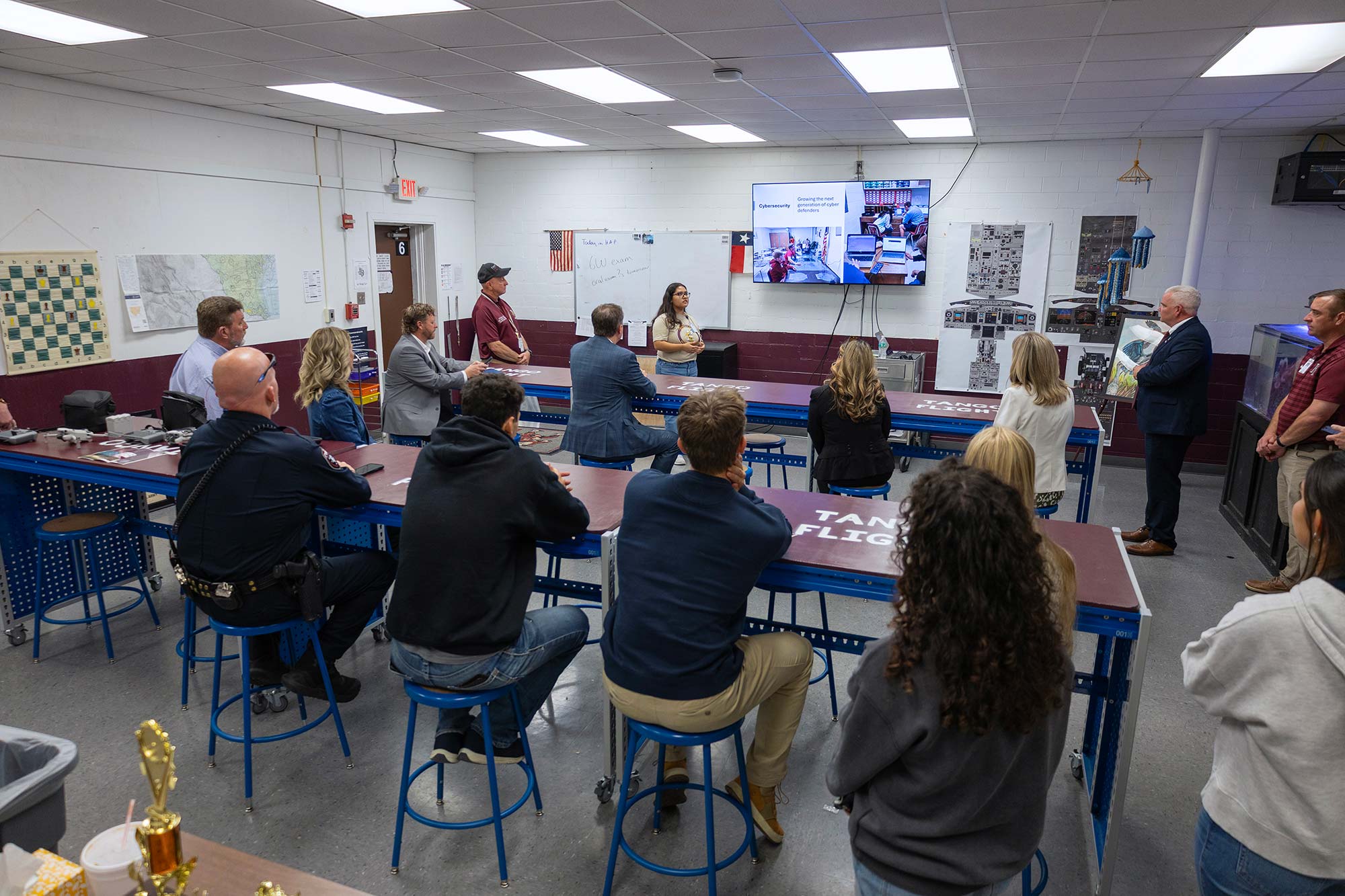 A group of adults sits and stands around tables in a classroom, listening to a woman speaking at the front. A screen behind her displays a video call, and various posters and maps decorate the walls.