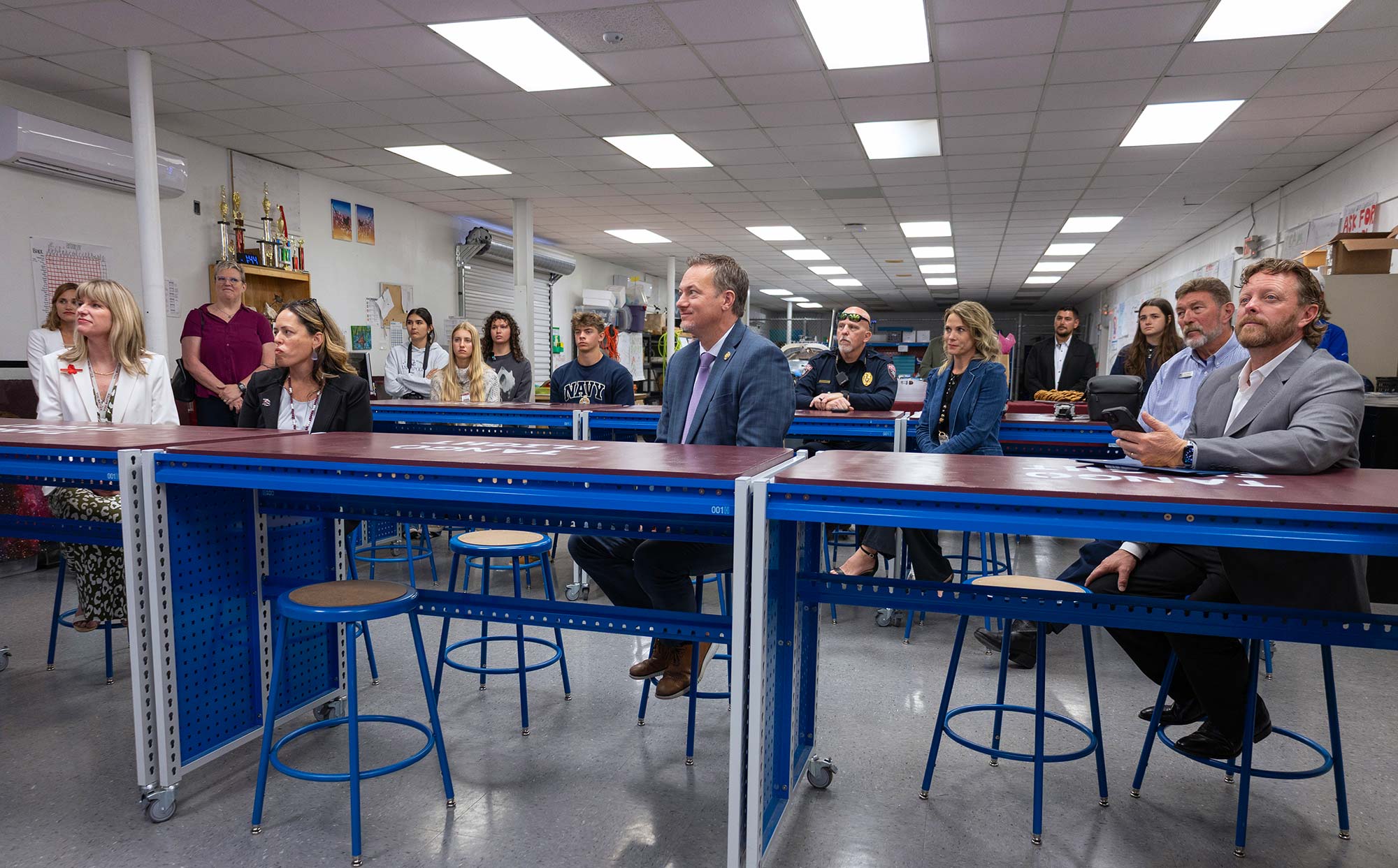 A group of adults sit attentively at blue tables in a classroom or workshop with tools and trophies on the shelves behind them, under a ceiling of fluorescent lights.