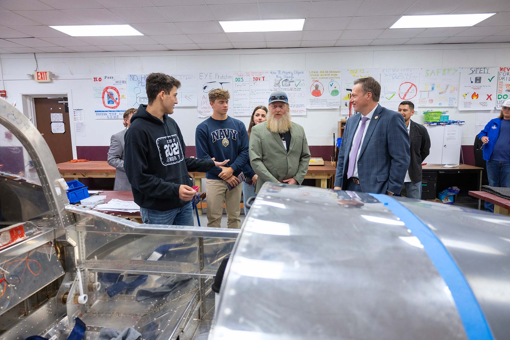 A group of students and adults stand around a partially assembled aircraft in a classroom, discussing the project. Posters and diagrams are visible on the walls in the background.