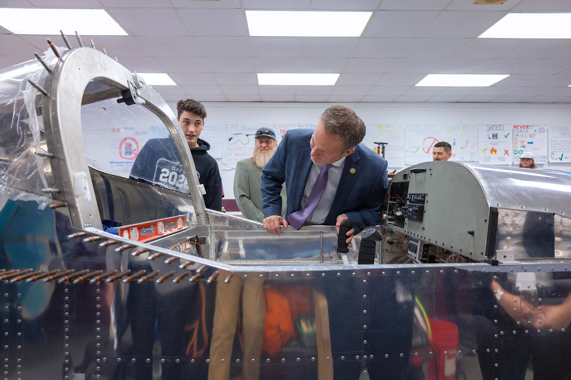 A man in a suit inspects the inside of an unfinished aircraft while a group of students and an older man watch in a classroom decorated with posters.
