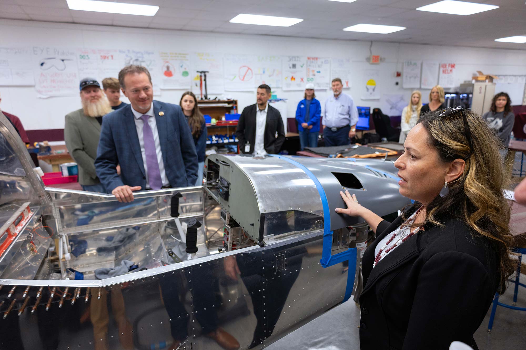 A woman explains an aircraft project to a group of people in a classroom. The group observes the partially assembled plane, while posters and diagrams are visible on the walls in the background.