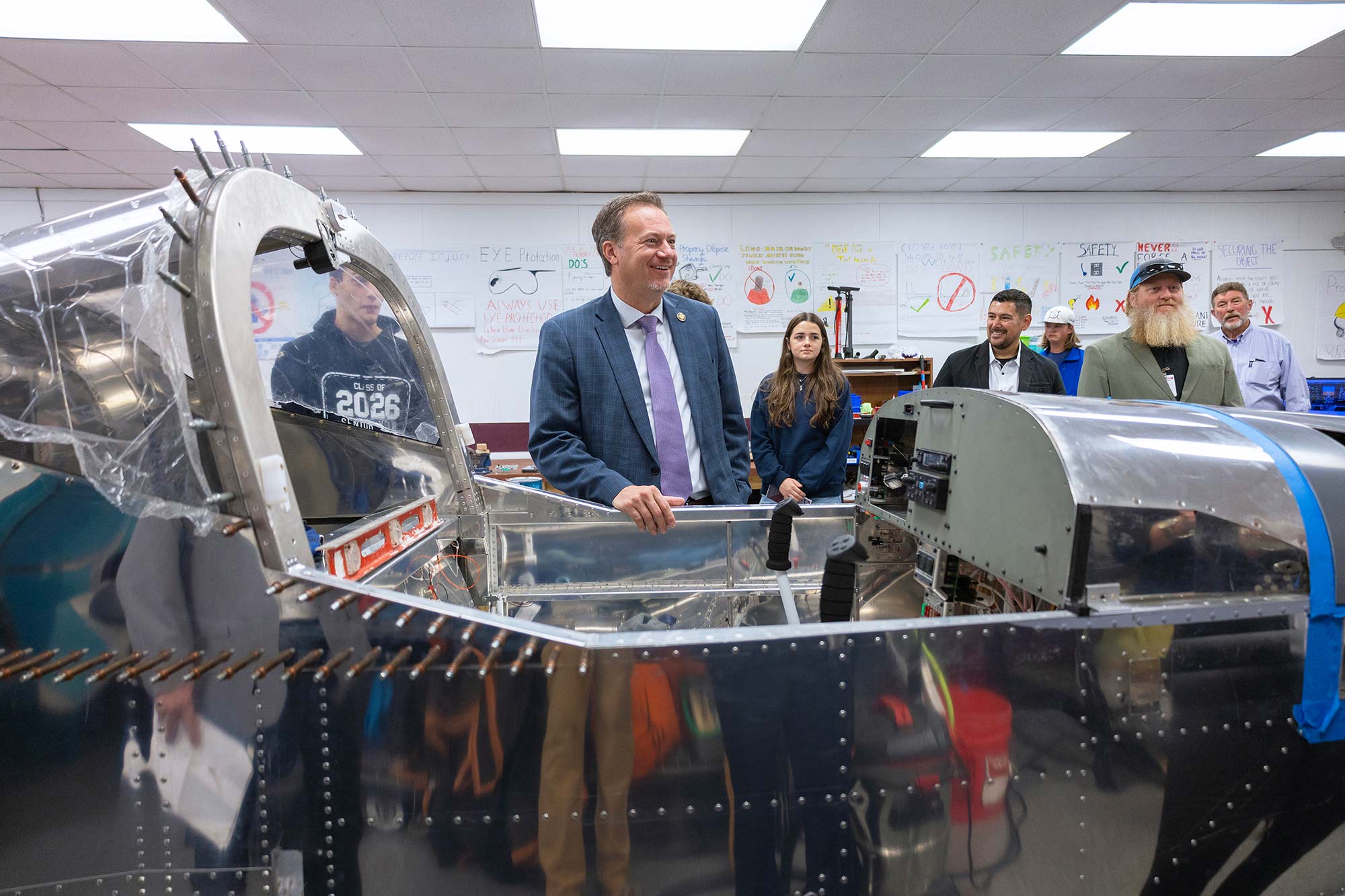 A group of people stand around a partially assembled aircraft in a classroom. A man in a suit smiles at the center, while others watch and converse. Educational posters and tools are visible in the background.