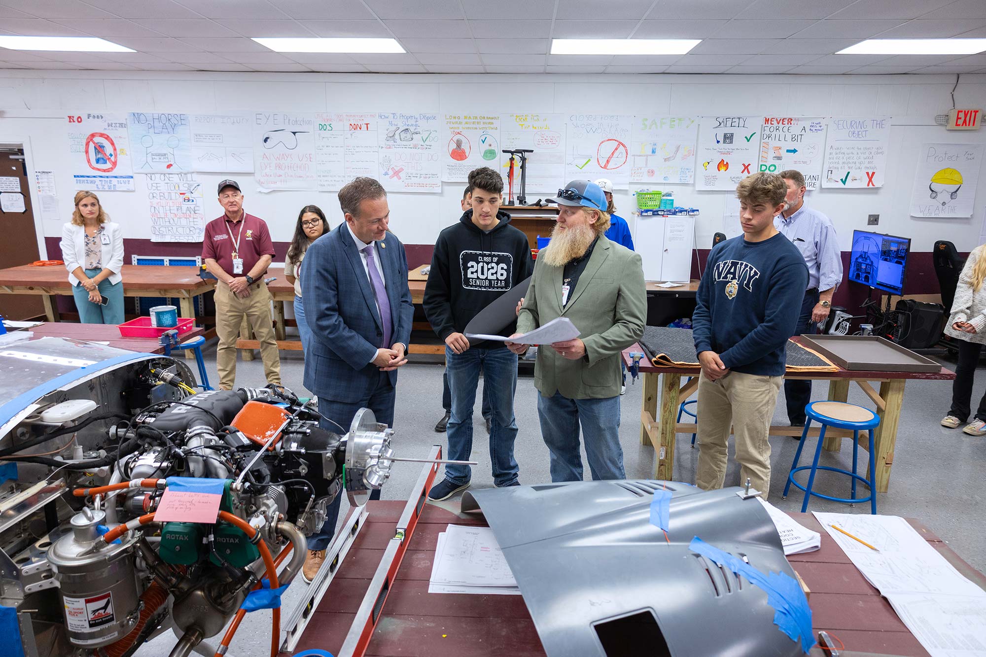 A group of people, including students and adults, stand in a classroom with posters on the walls, reviewing papers beside a partially assembled vehicle and various mechanical parts on tables.