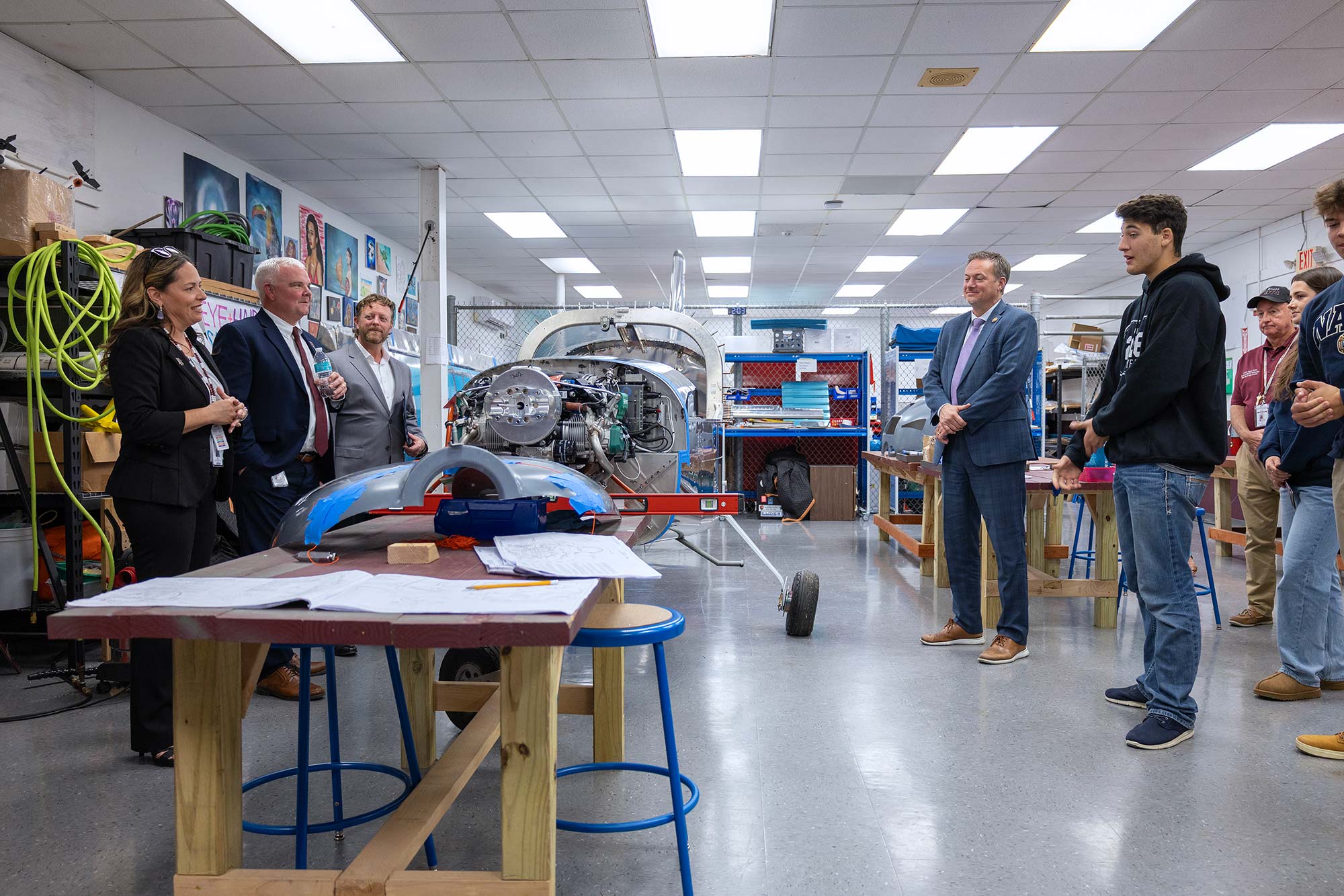 A group of people, including students and adults in business attire, stand in a workshop surrounded by tools and machinery, discussing a partially assembled vehicle on a table in the center of the room.