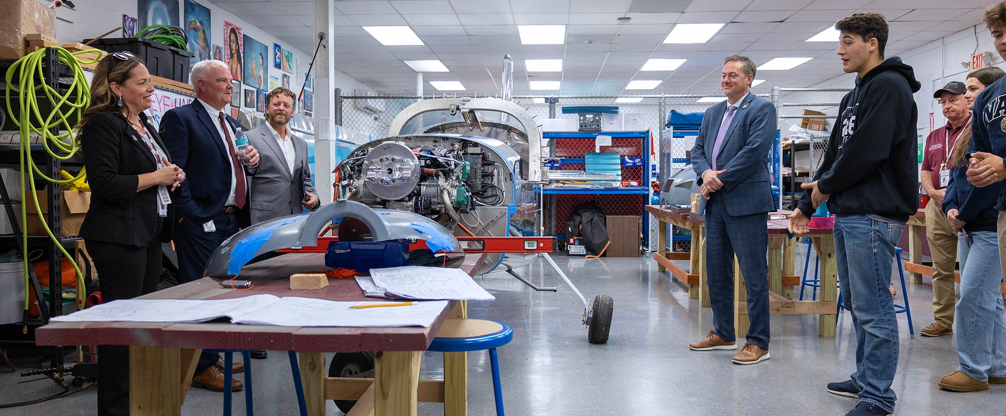 A group of people stand in a workshop near a partially assembled small airplane, with tools, equipment, and blueprints visible around the room. Some are listening while one person appears to be speaking.