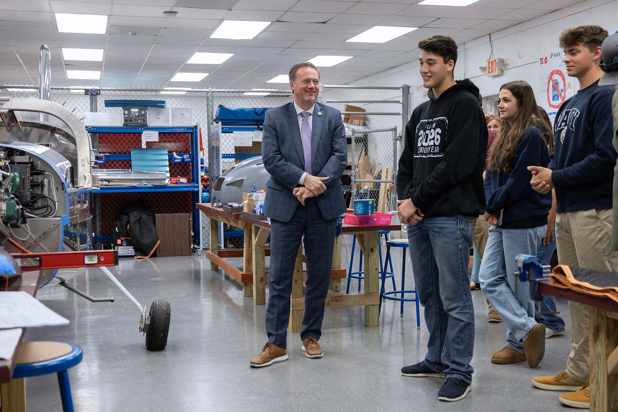 A man in a suit stands smiling with several casually dressed students in a workshop or classroom, surrounded by workbenches, tools, and a partially assembled aircraft.