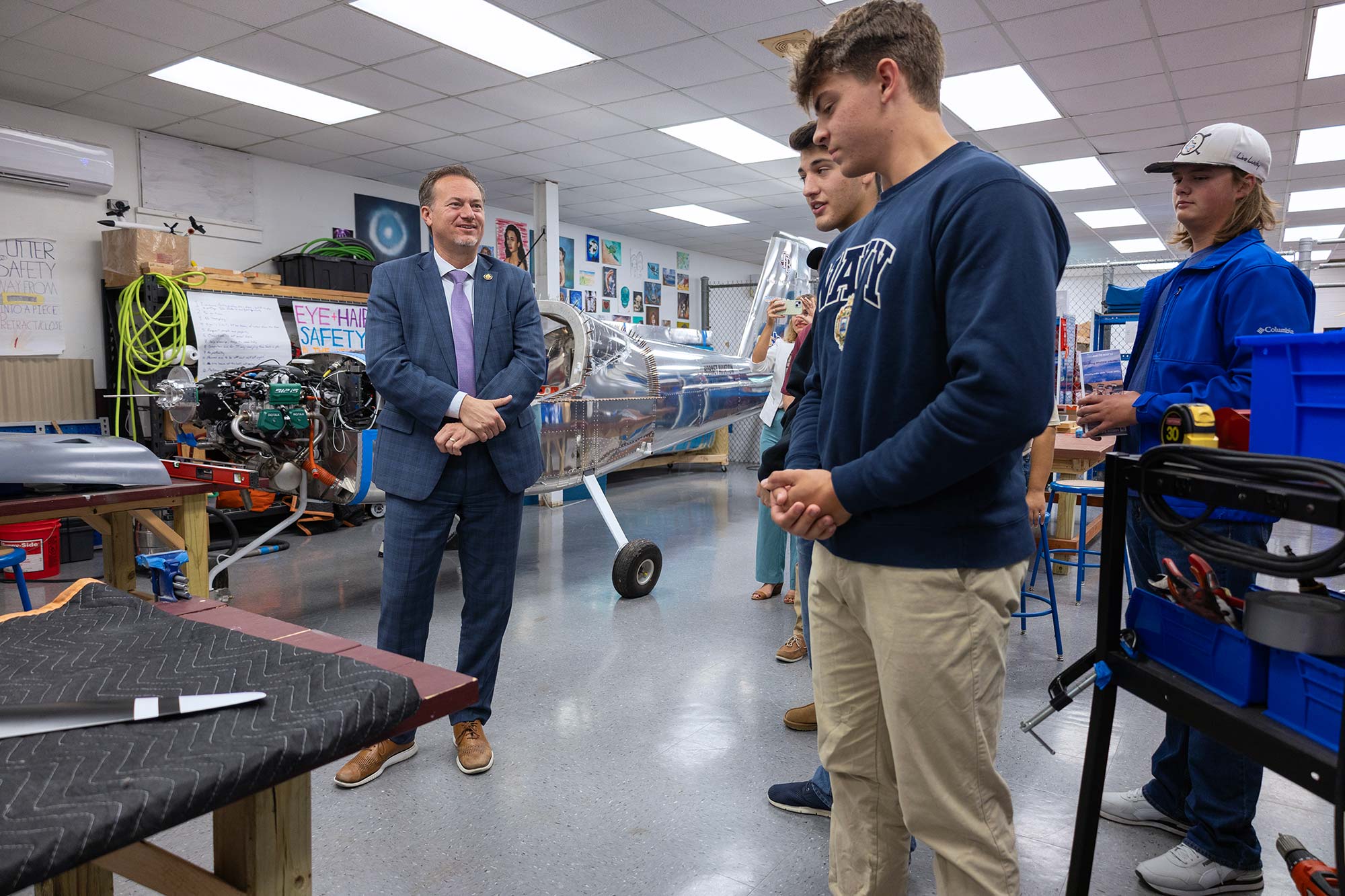A group of students talks with a man in a suit inside a workshop with a small airplane under construction. The room has tools, equipment, and safety posters visible in the background.