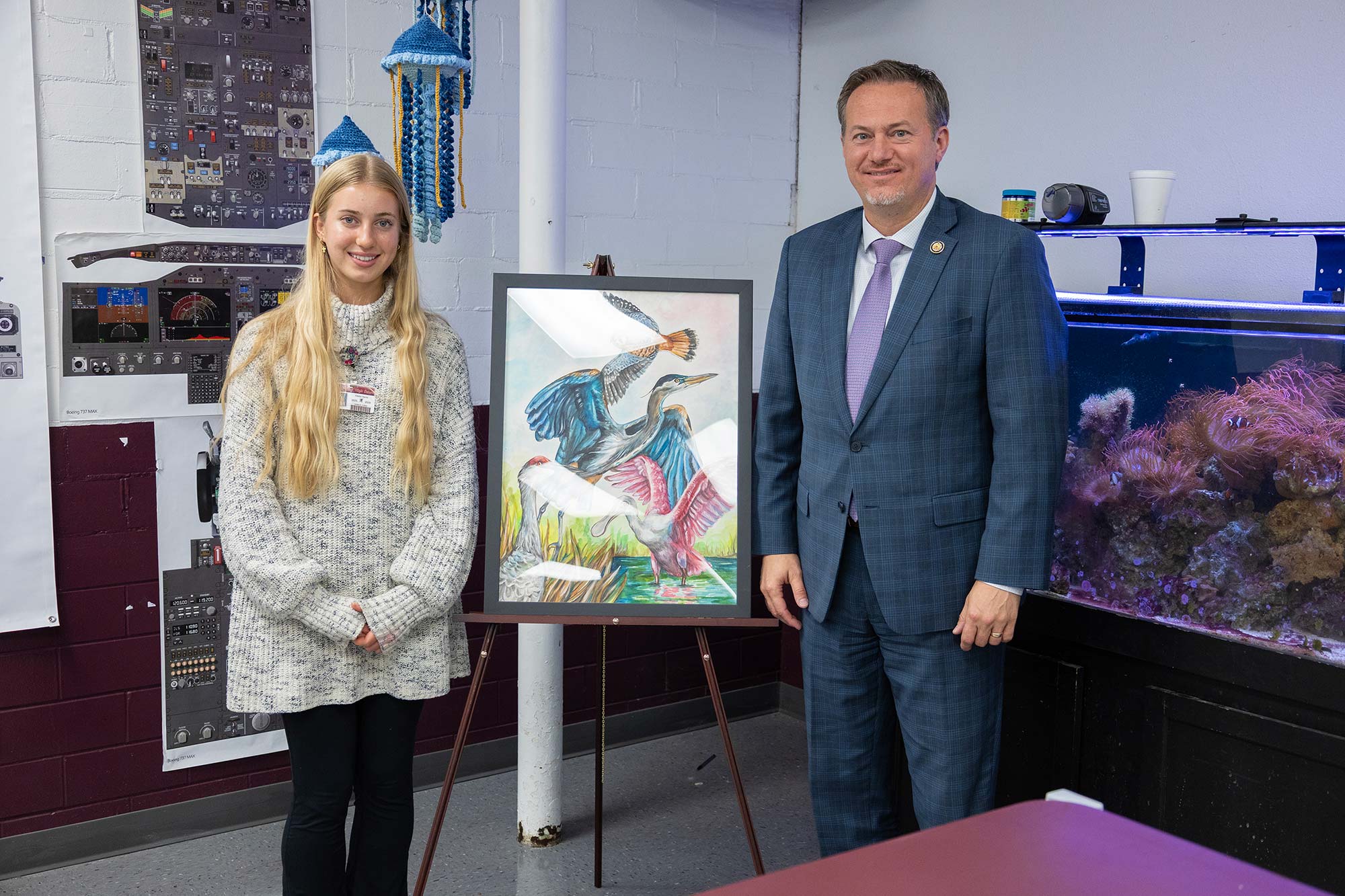 A young woman and a man in a suit stand beside a colorful painting of birds on an easel in a classroom setting with posters and an aquarium in the background.