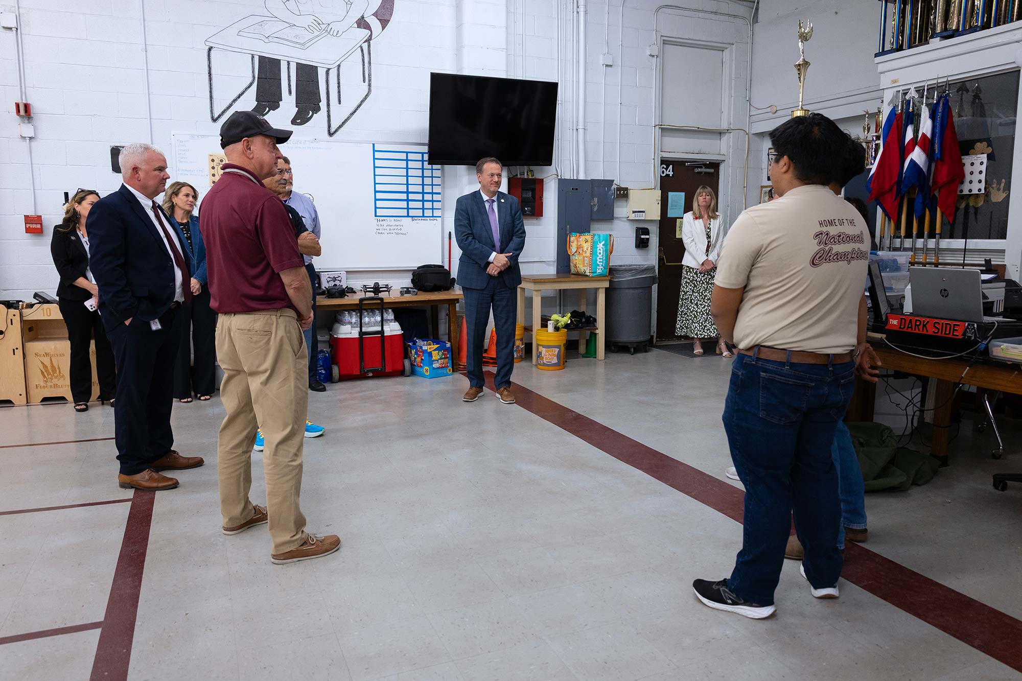 A group of people, including men in business attire and casual clothes, stand in a classroom or workshop having a discussion. Equipment, flags, and a TV are visible in the background.