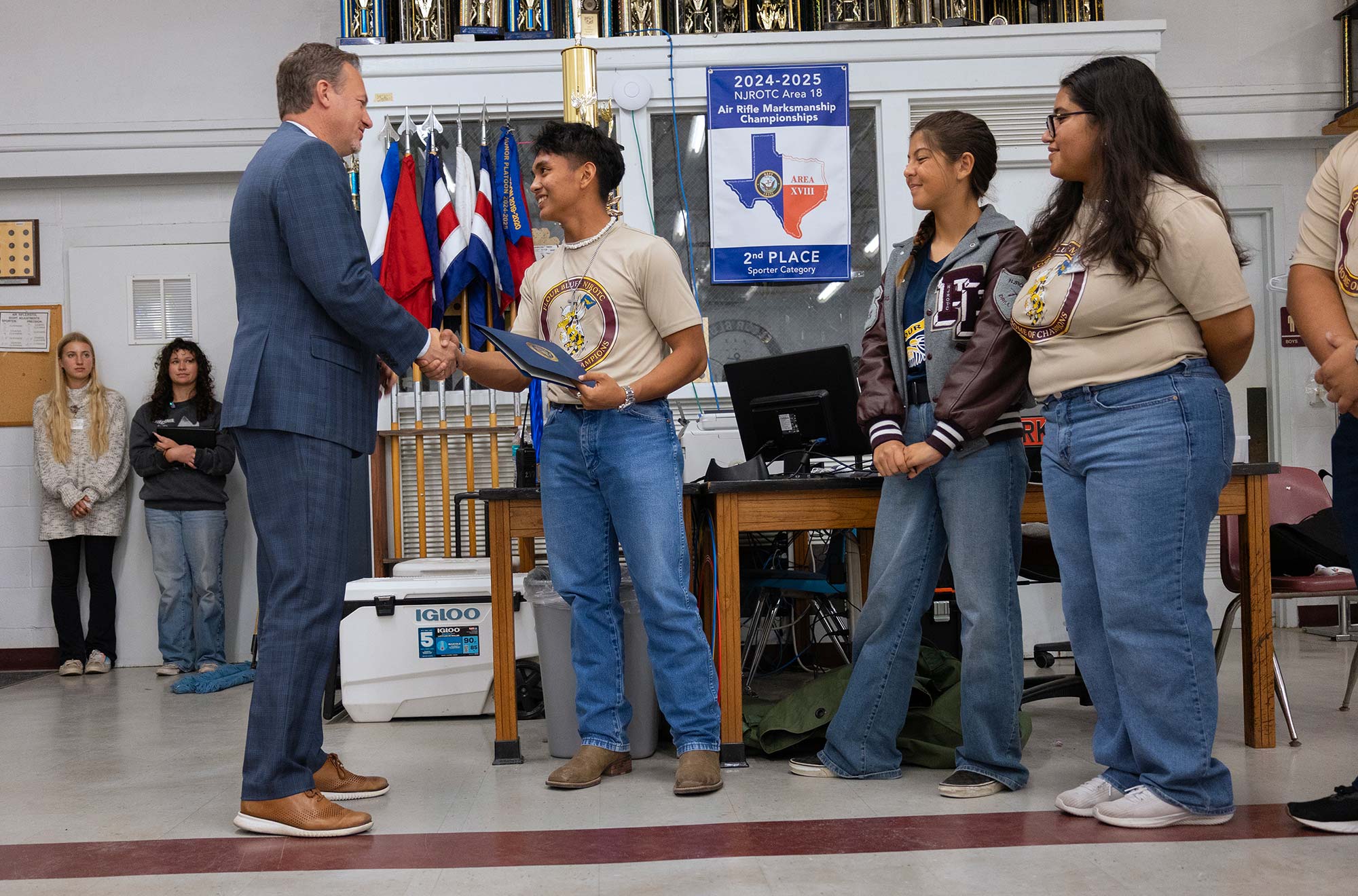 A man in a suit shakes hands with a smiling student holding a certificate, while three other students stand nearby, all wearing matching shirts. Others watch in the background in a classroom decorated with flags and trophies.